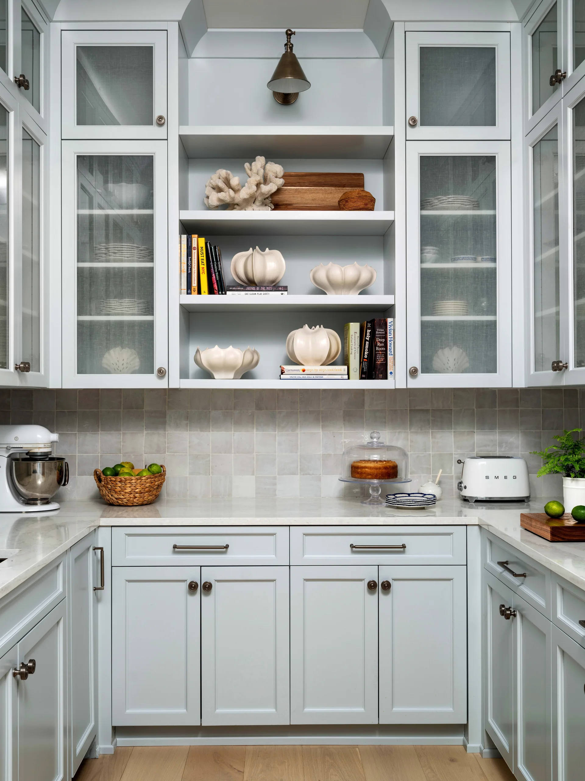 Light blue kitchen cabinets flank central open shelving decorated with white sculptural vases, set above a grey square tile backsplash and white countertop.