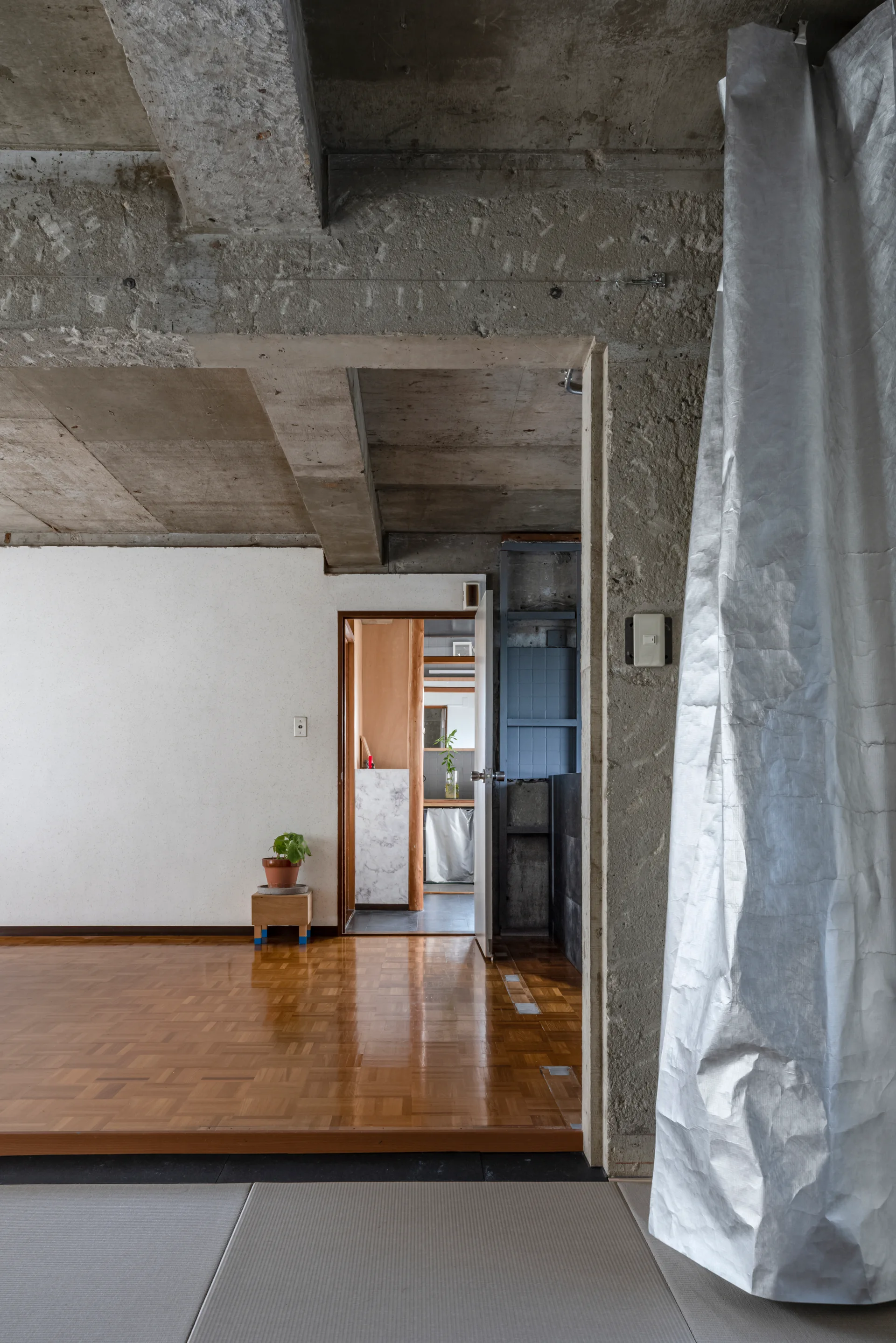 A minimalist room features an exposed raw concrete ceiling and polished parquet wood flooring, with an open doorway leading to another space.