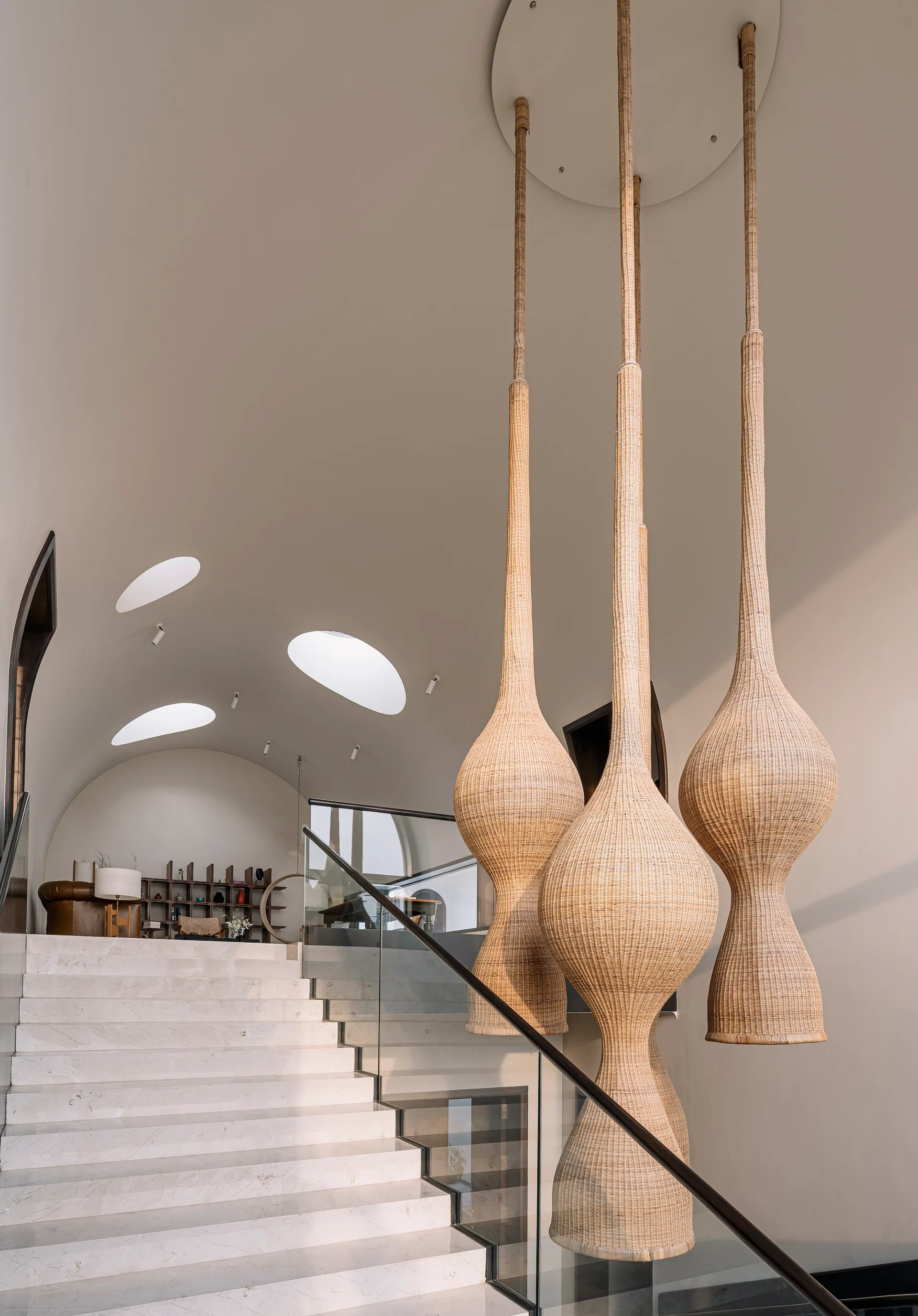 Oversized woven wicker pendant lights hang beside a white marble staircase with a glass railing under a vaulted ceiling with elliptical skylights.