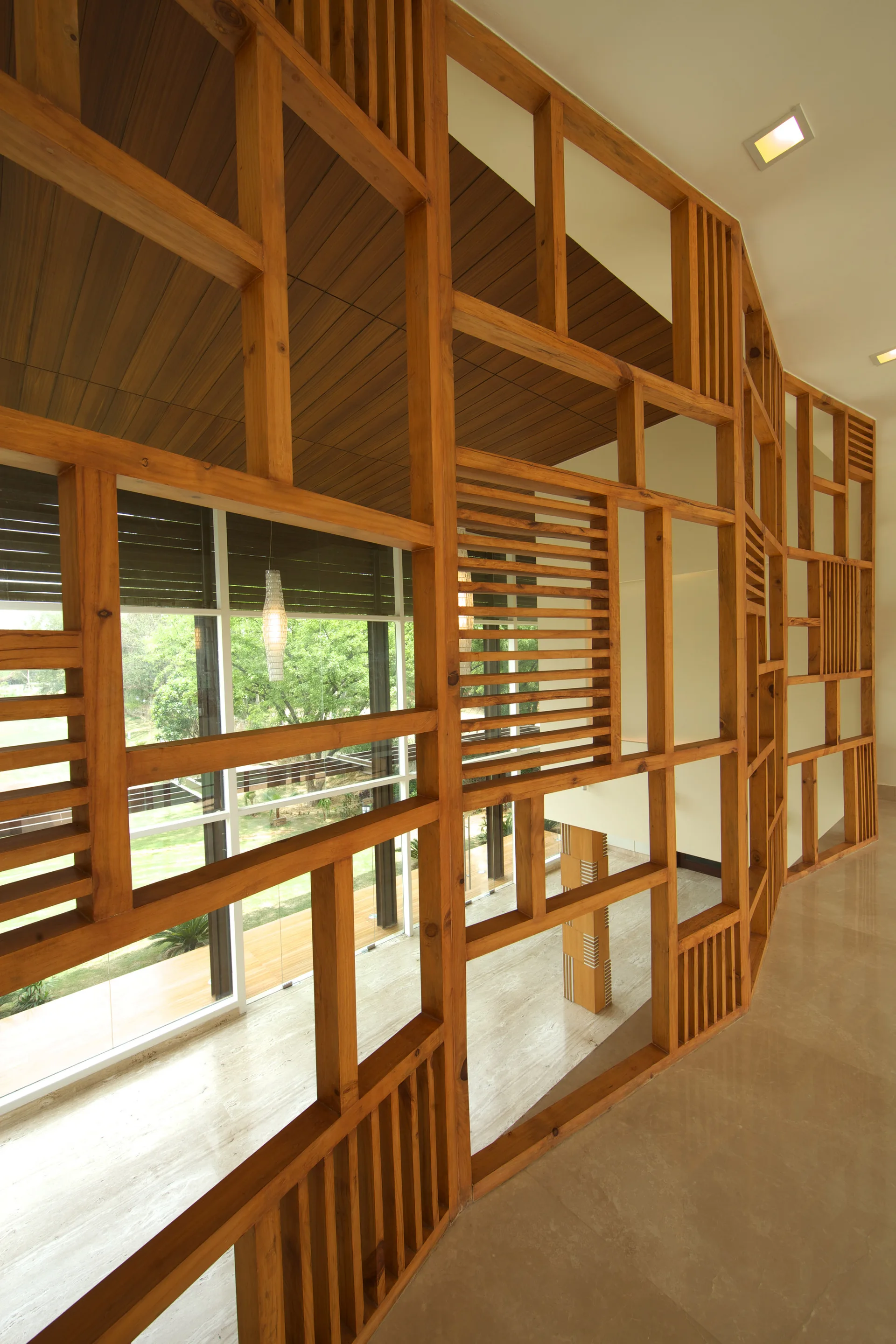 A tall wooden lattice screen with a geometric grid pattern stands along a polished hallway next to large windows overlooking green trees.
