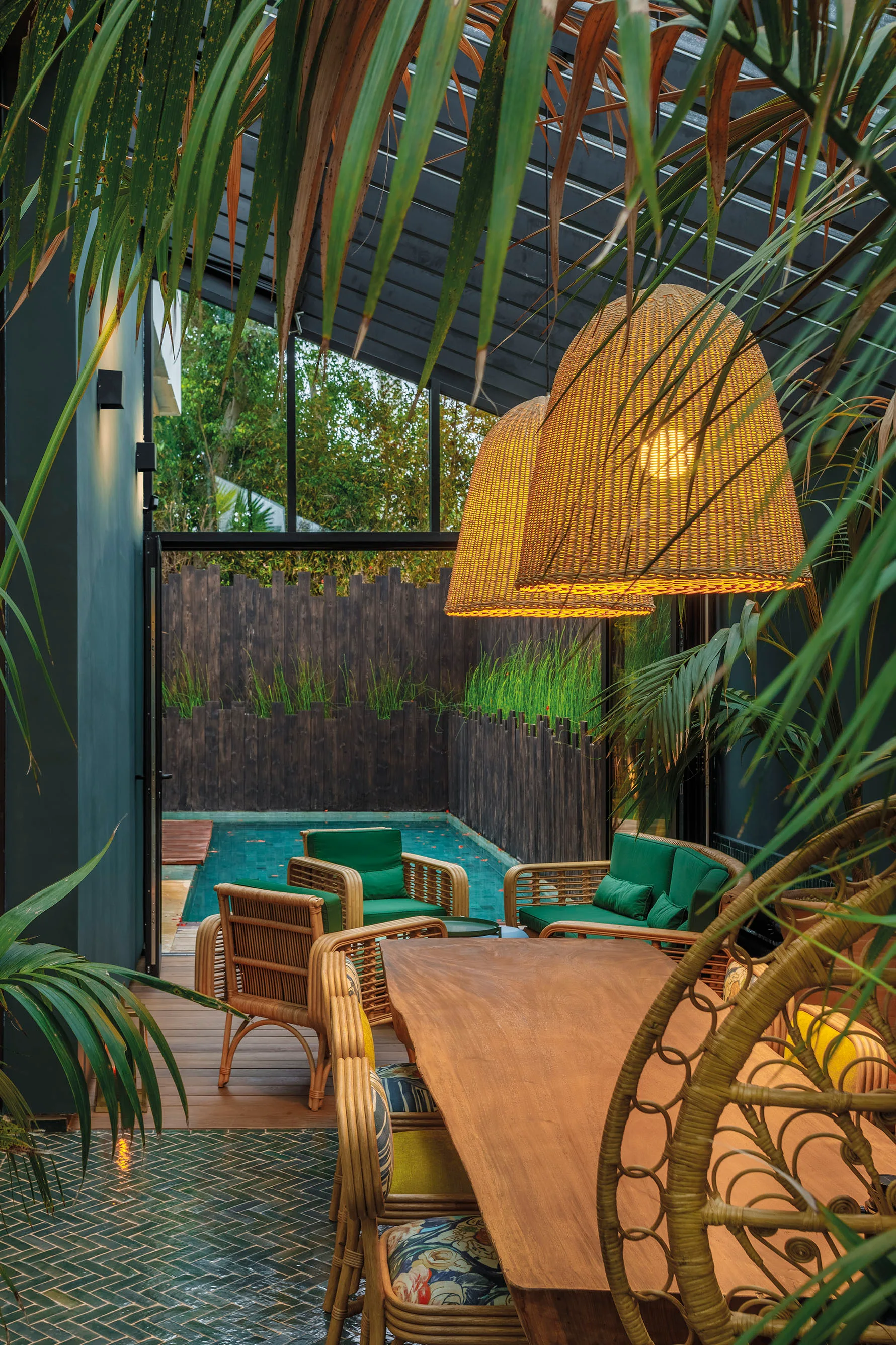 A tropical patio features a wooden dining table, rattan chairs with green cushions, and large woven pendant lights overlooking a swimming pool.