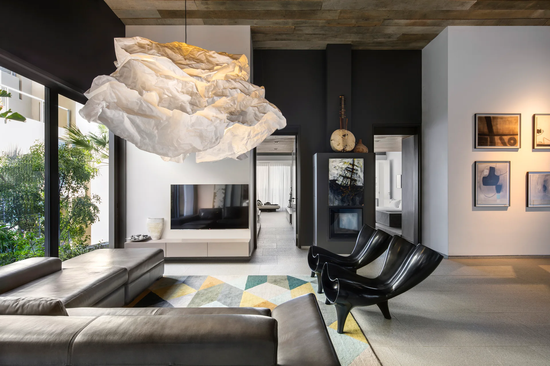 A modern living room features a large crinkled paper pendant light, grey leather sectional, black sculptural chairs, and a wood plank ceiling.