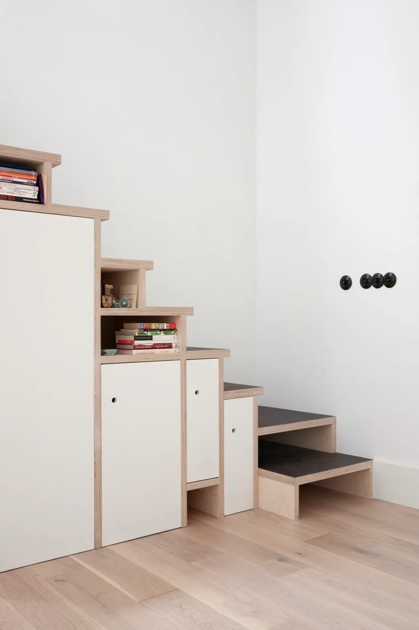 Stepped plywood staircase featuring integrated white laminate cabinets, open storage shelves with books, and circular black wall hooks.