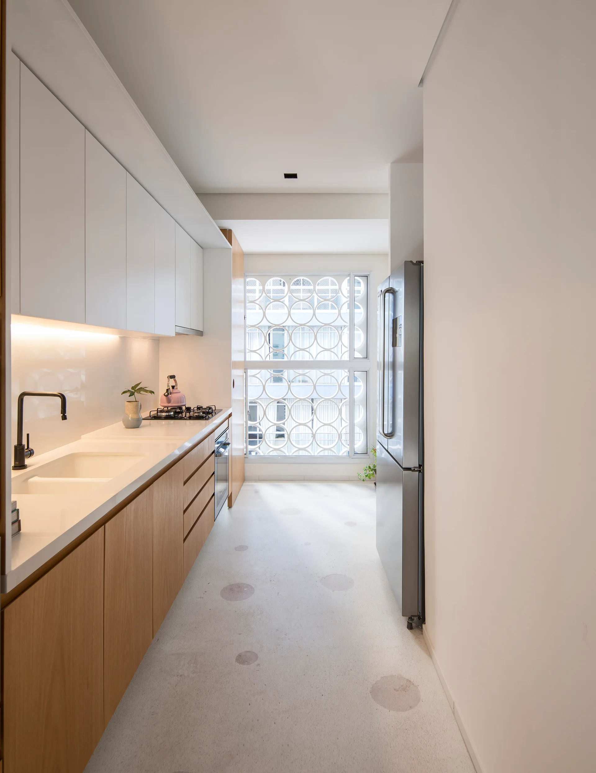 A modern galley kitchen featuring white upper and wood lower cabinets, a stainless steel refrigerator, and a window with a circular lattice screen.