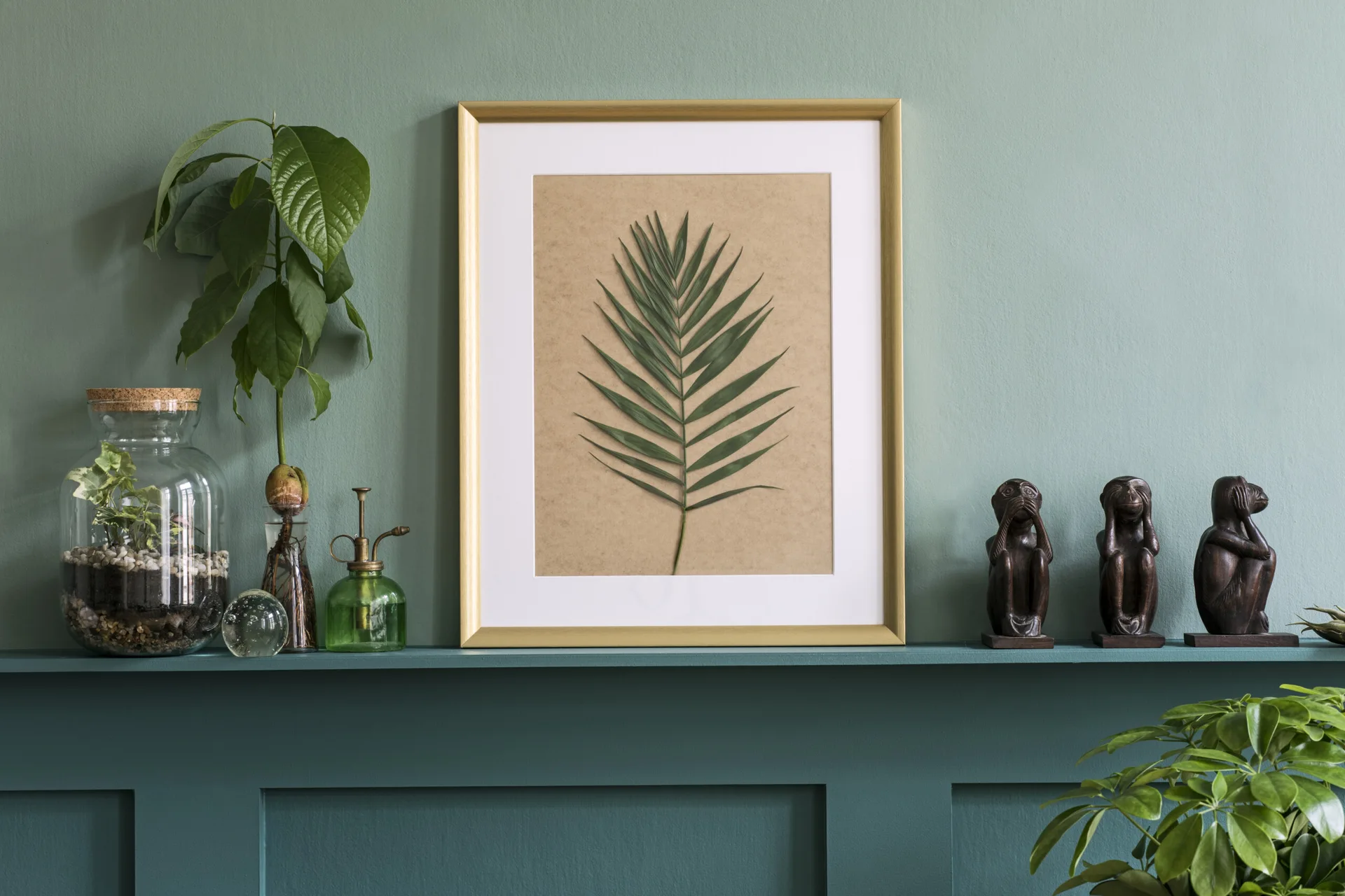 A gold-framed botanical palm leaf print sits on a dark teal paneled shelf alongside glass terrariums and small wooden figurines.