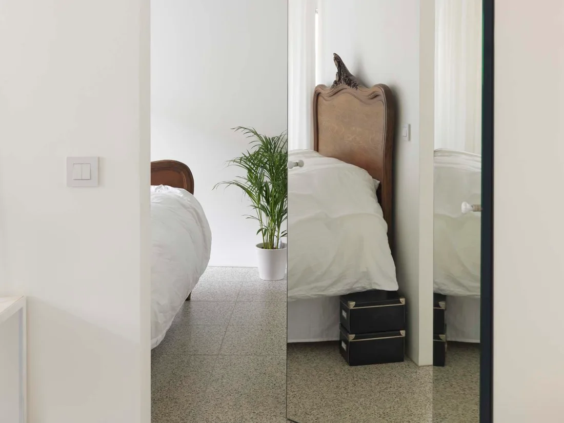 A bedroom viewed through a vertical mirror features an antique wooden headboard, white bedding, a potted palm plant, and a speckled terrazzo floor.