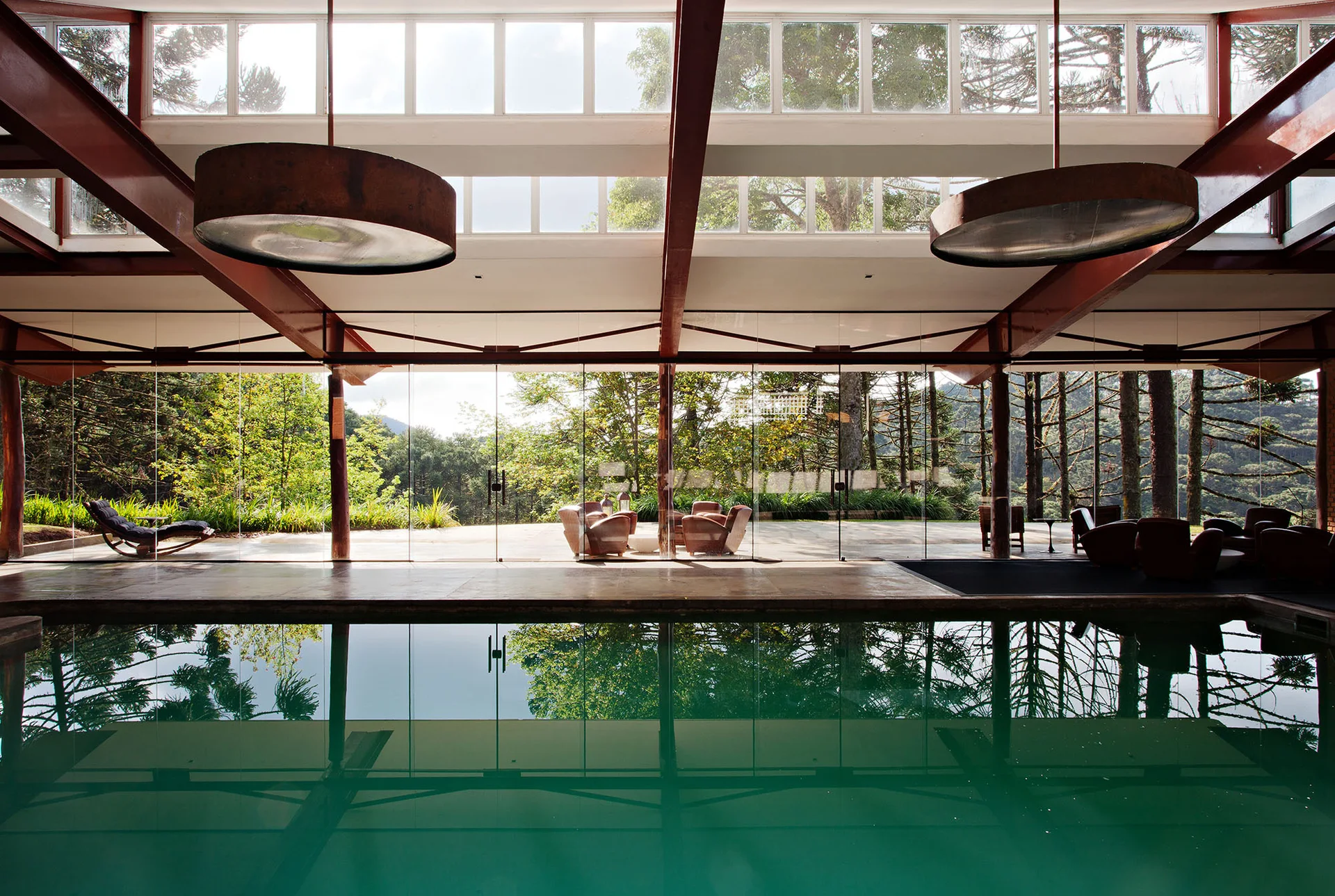 An indoor pool reflects the surrounding forest through expansive floor-to-ceiling glass walls beneath a red steel beam ceiling.
