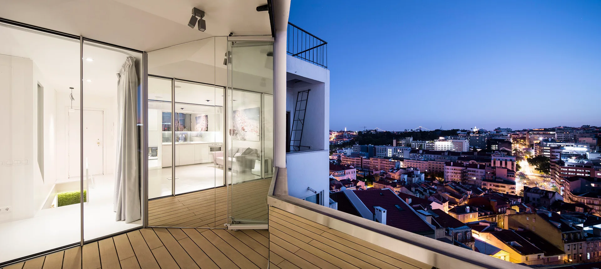 A modern high-rise balcony features wood-toned decking, a folding glass wall enclosure, and an expansive evening view of a city skyline.