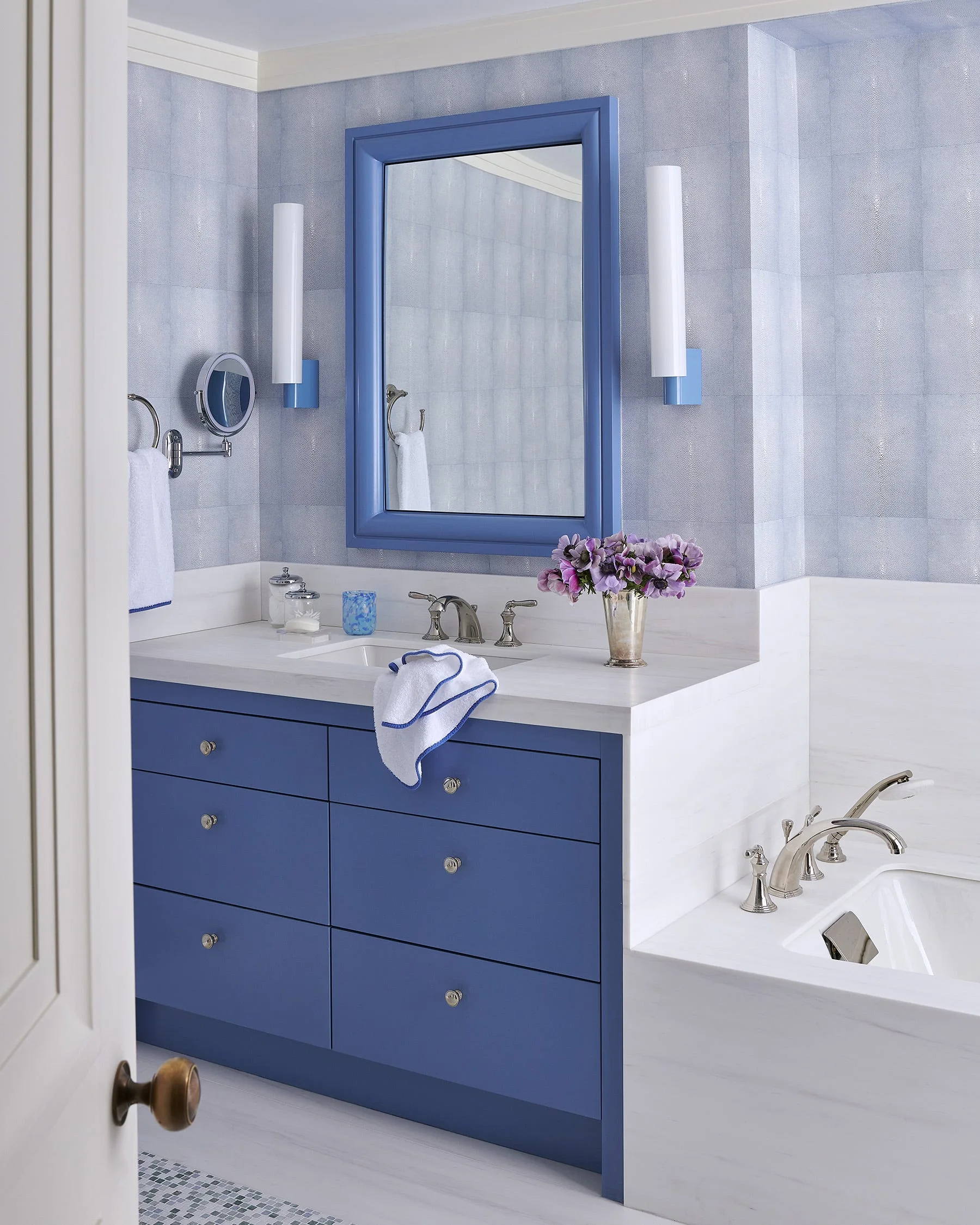 A blue bathroom vanity with a white stone countertop sits beneath a matching blue framed mirror and modern vertical wall sconces.