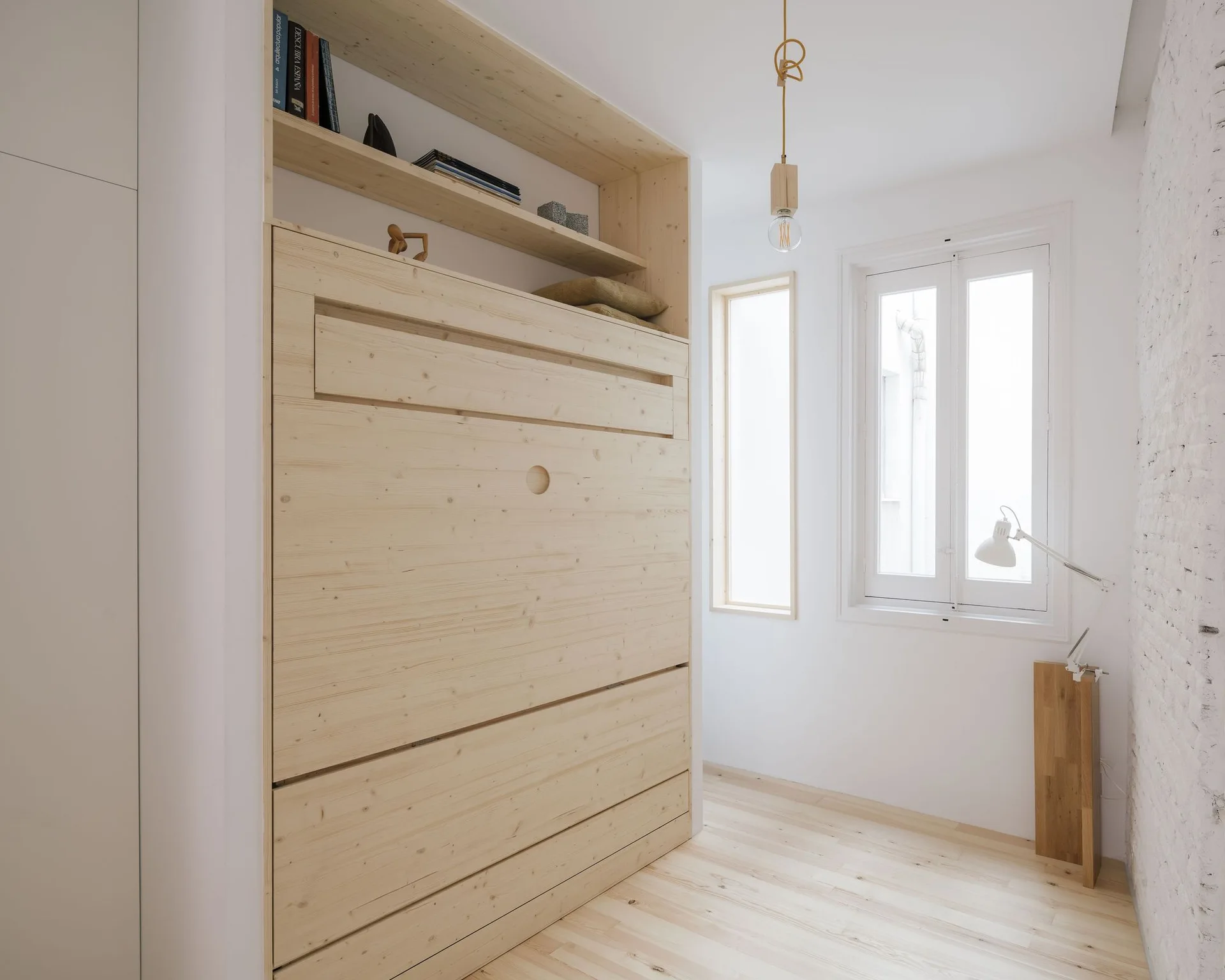A minimalist white bedroom features a light wood wall bed with top shelving, light wood floors, and a textured white brick wall.