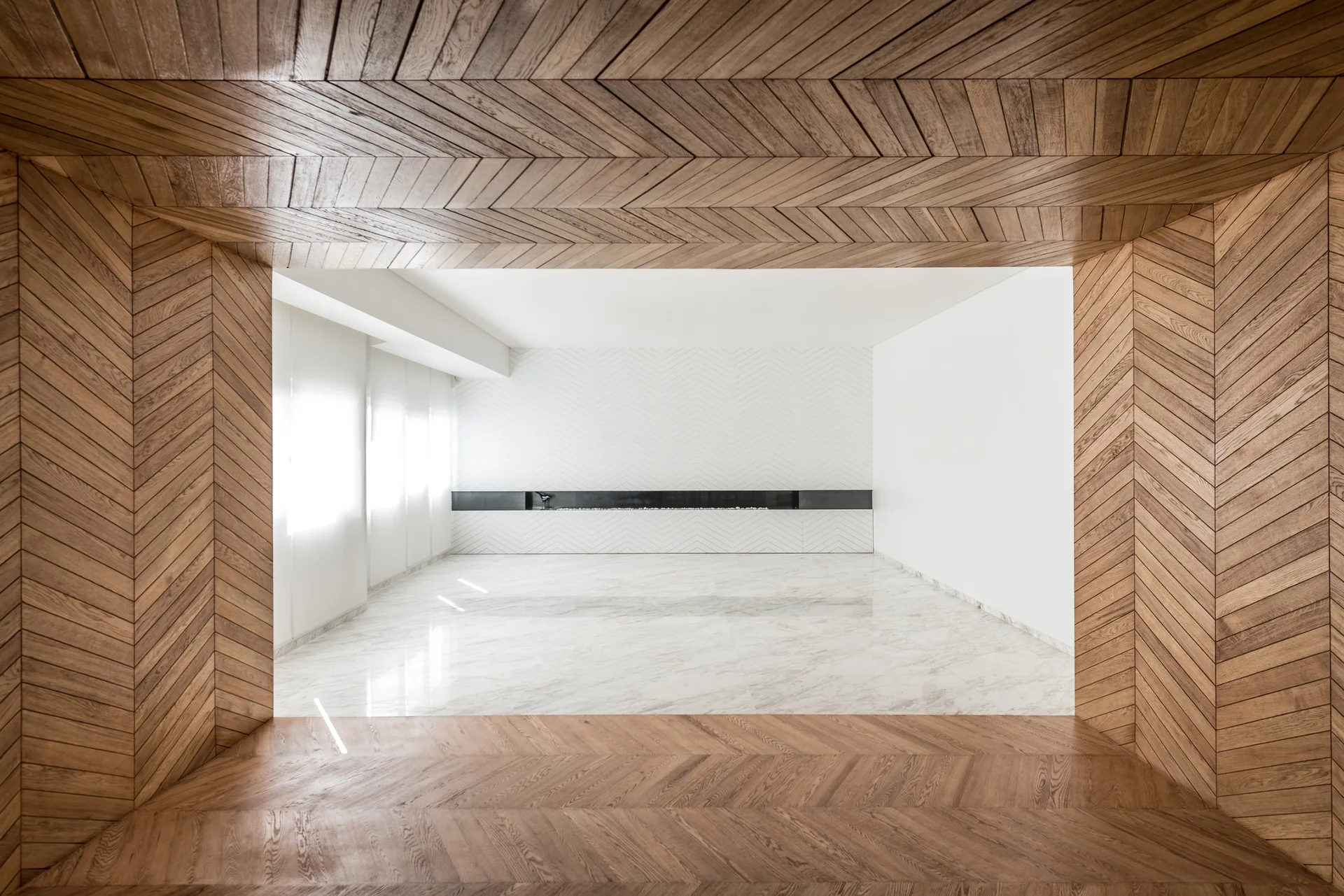 A minimalist living room with a wood herringbone paneled ceiling and walls, white marble flooring, and a long black linear fireplace.