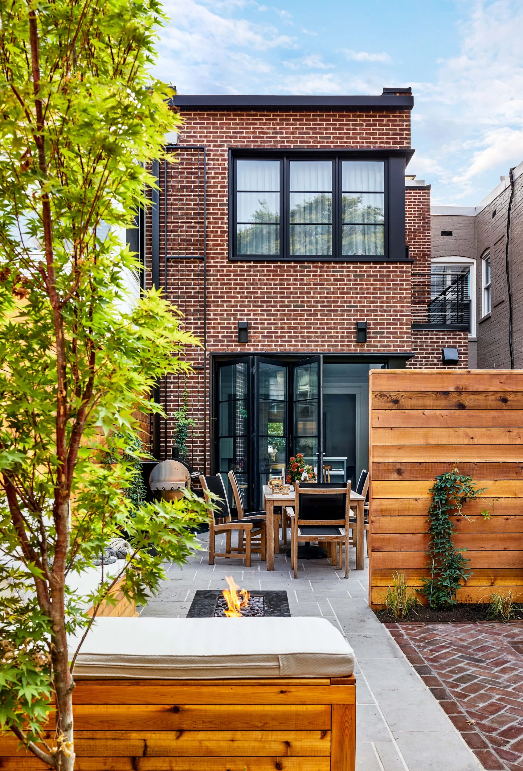 A backyard patio with a red brick exterior, featuring a wood dining table, a horizontal plank privacy fence, and a modern fire pit.