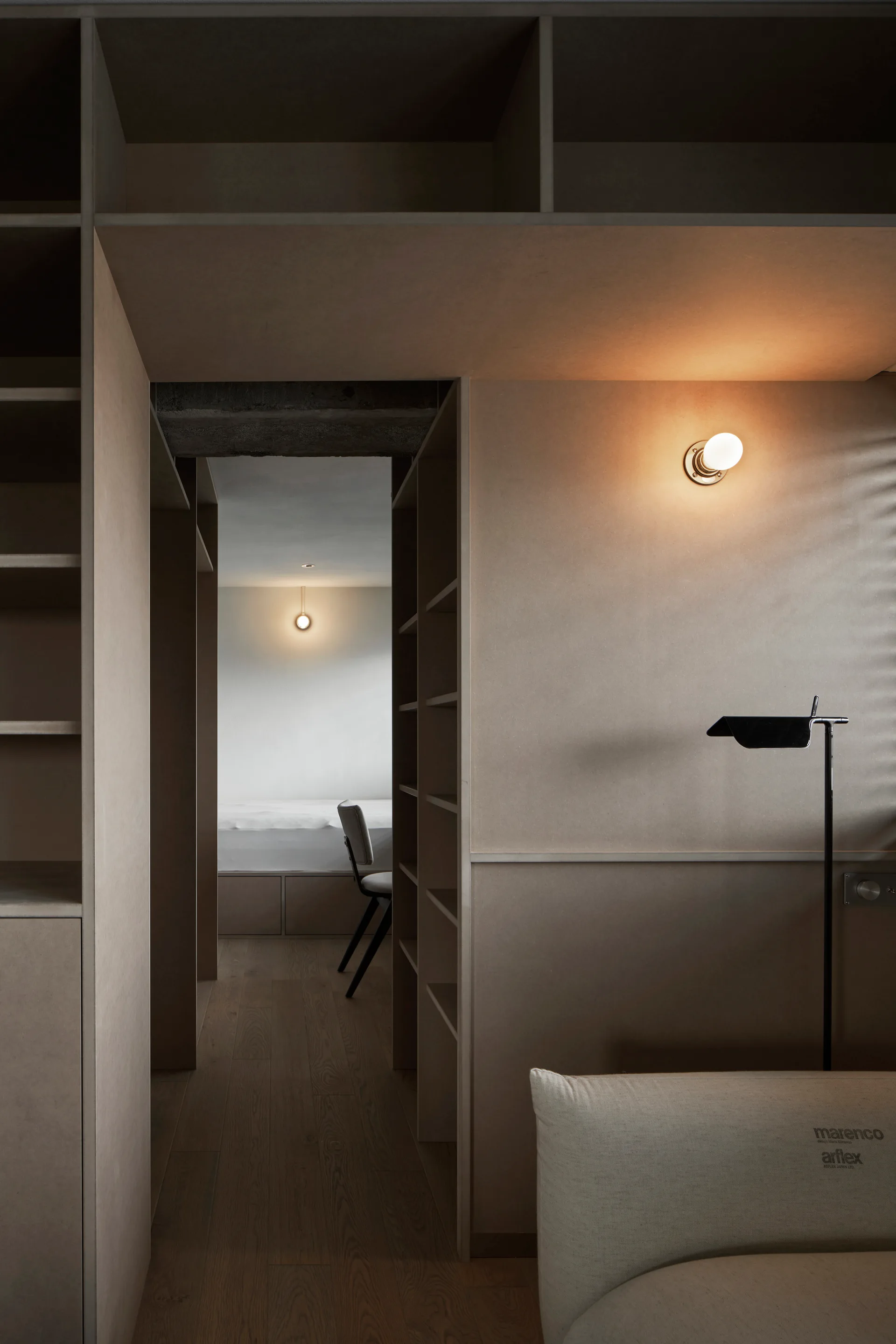 A modern bedroom view through a custom beige wooden partition featuring built-in shelving, a black floor lamp, and glowing wall sconces.