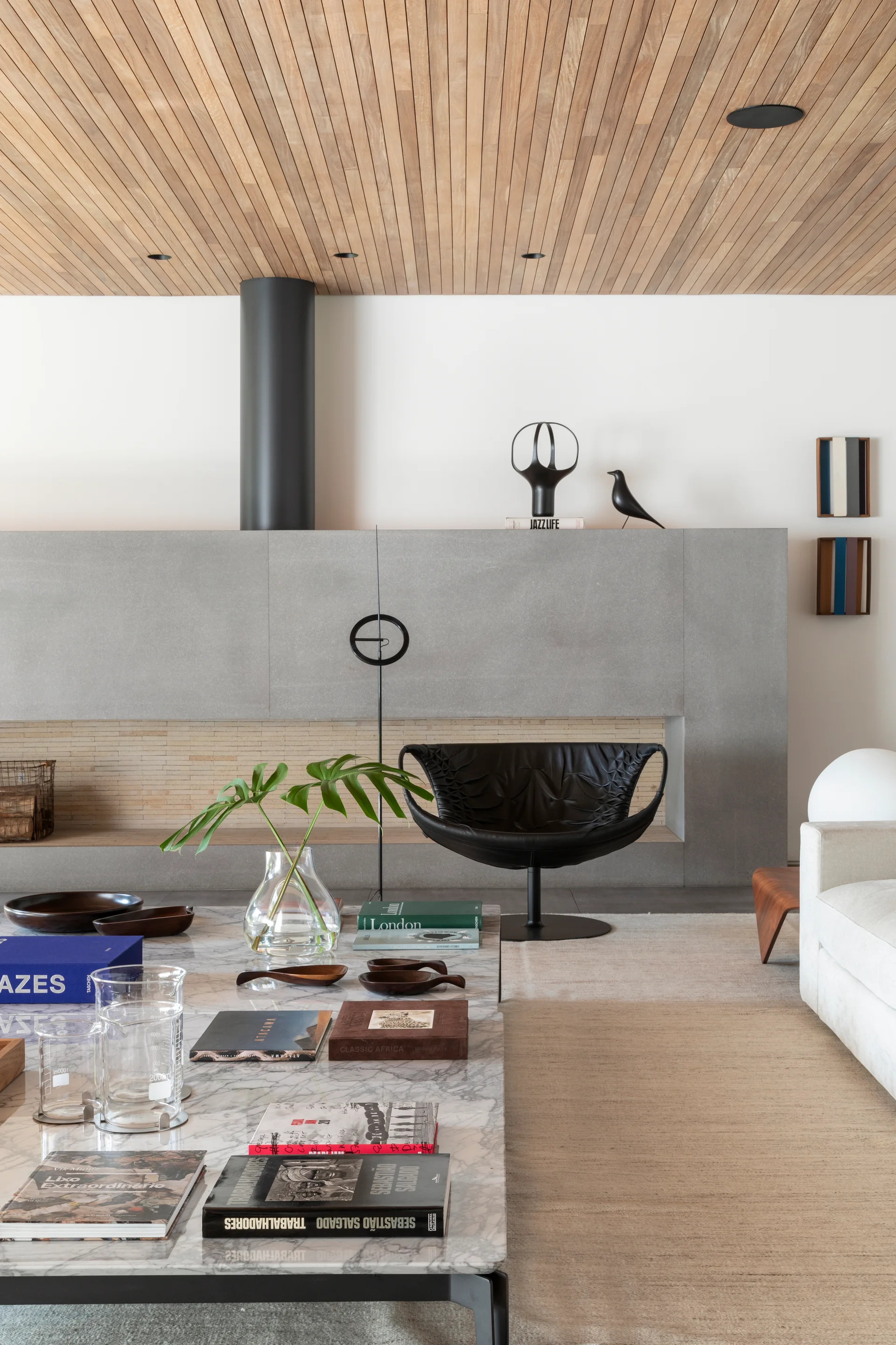 A modern living room features a wood-slatted ceiling, a grey concrete fireplace, a black leather swivel chair, and a marble coffee table styled with books.