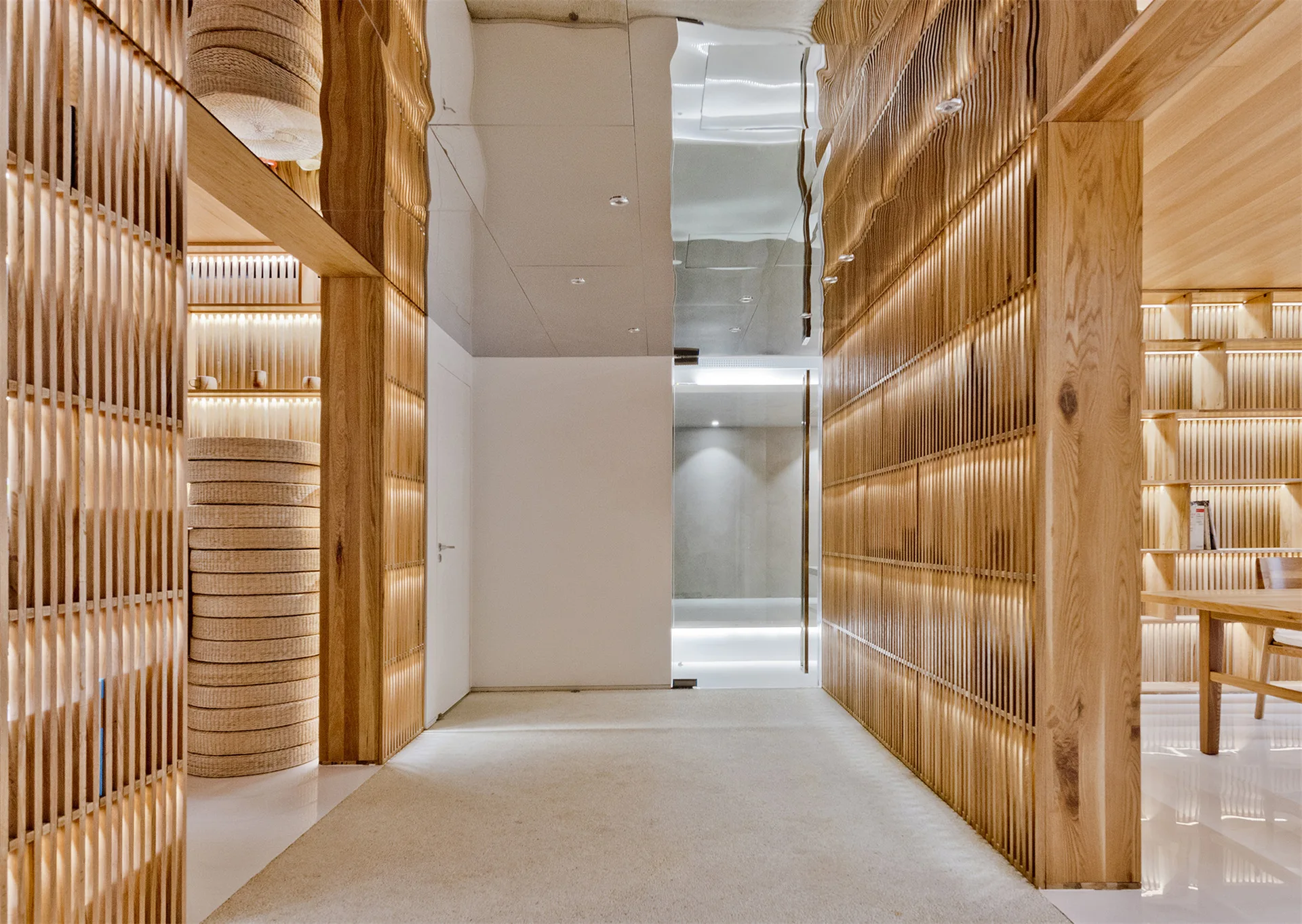 A modern hallway features vertical slatted wood walls with built-in warm lighting, a reflective mirrored ceiling, and neutral carpeted flooring.