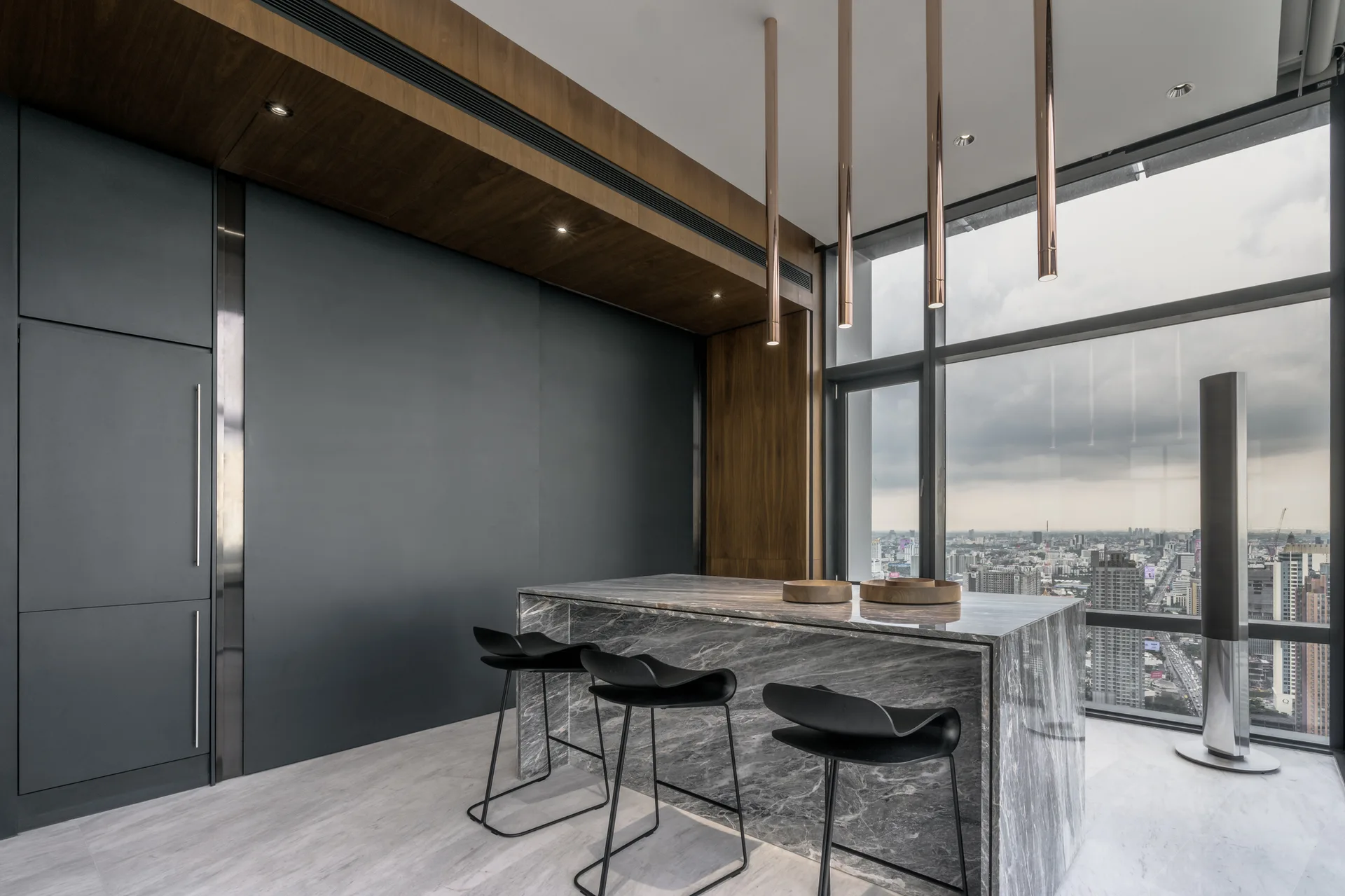 A modern kitchen area features a grey marble island with black stools, dark matte cabinetry, and copper pendant lights.