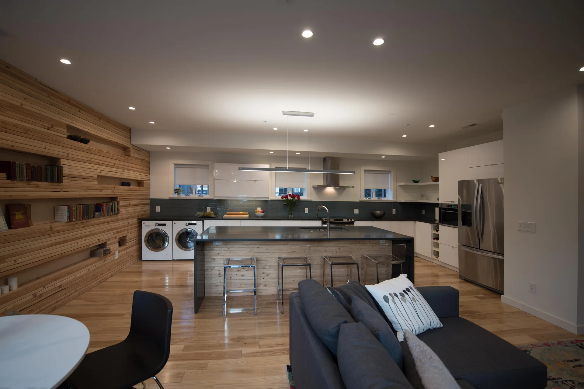 An open-concept kitchen features a horizontal wood slat accent wall, integrated laundry units under white cabinetry, and a central island with acrylic stools.