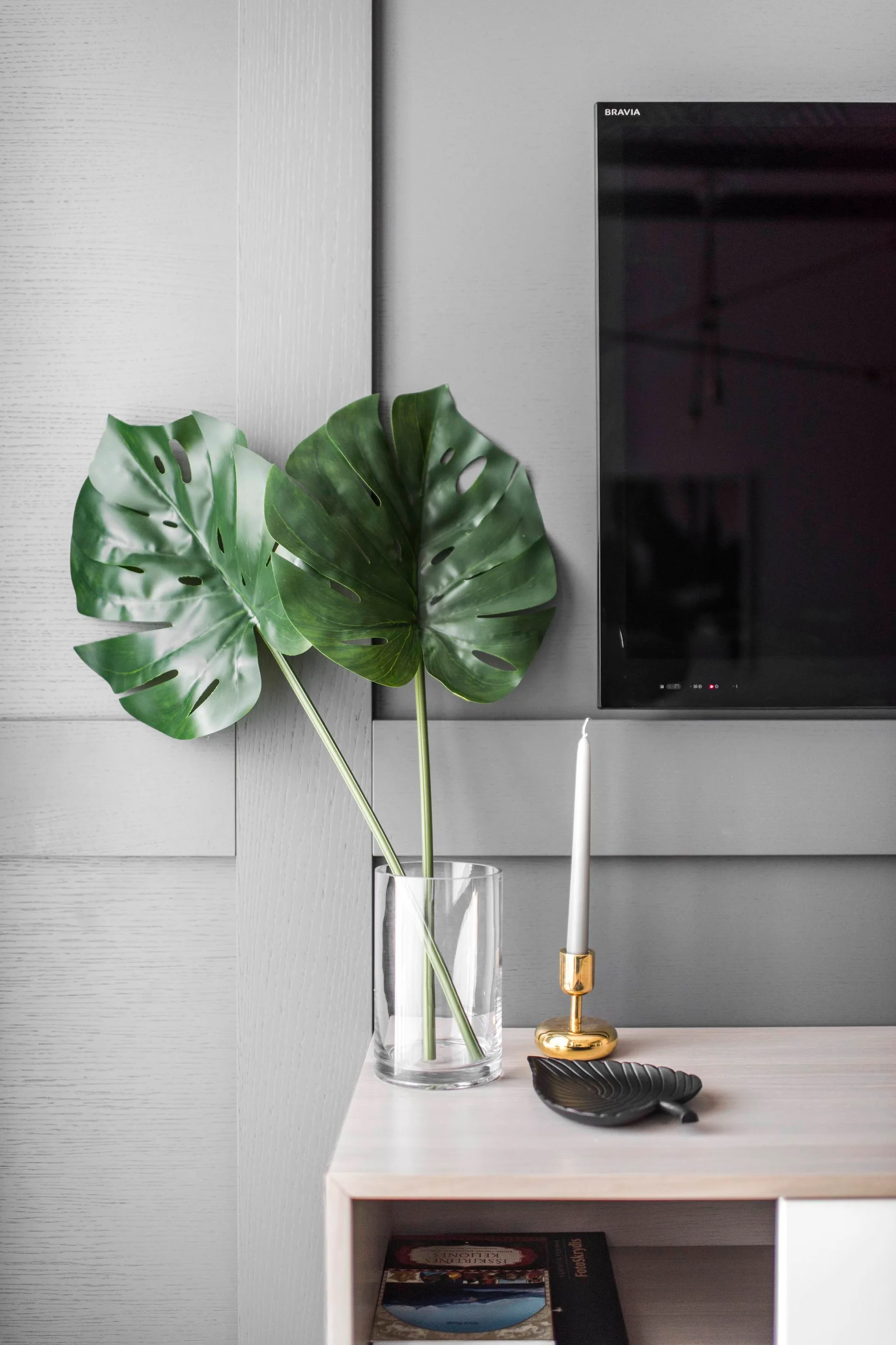 A light wood console table features a clear vase with monstera leaves, a gold candle holder, and a black dish against a grey paneled wall.