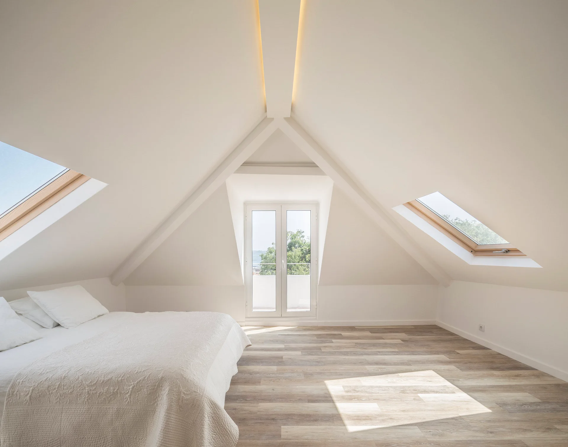 A minimalist attic bedroom featuring a white bed, sloped ceilings with skylights, and a central glass balcony door over light wood flooring.