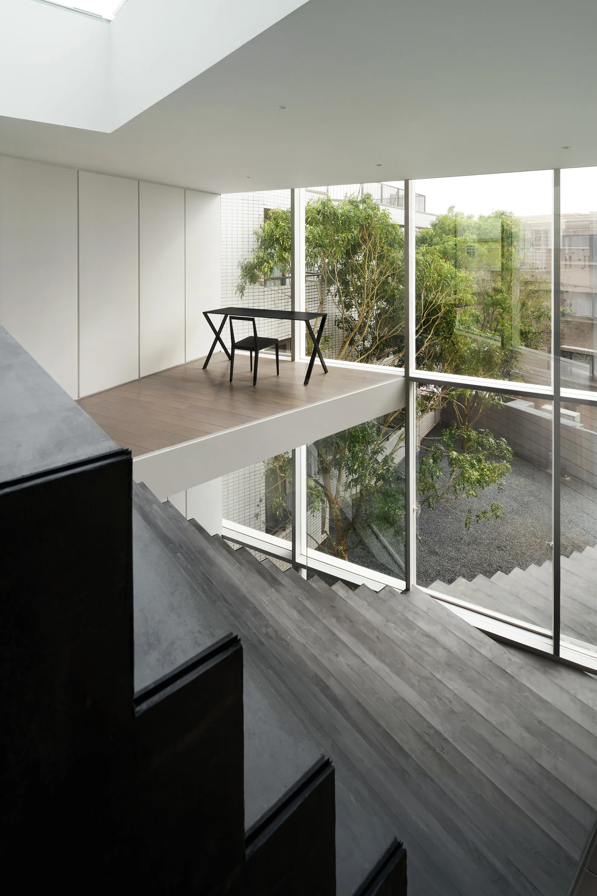 A dark concrete staircase leads up toward a wooden mezzanine platform featuring a minimalist black desk and chair in front of a giant glass window.