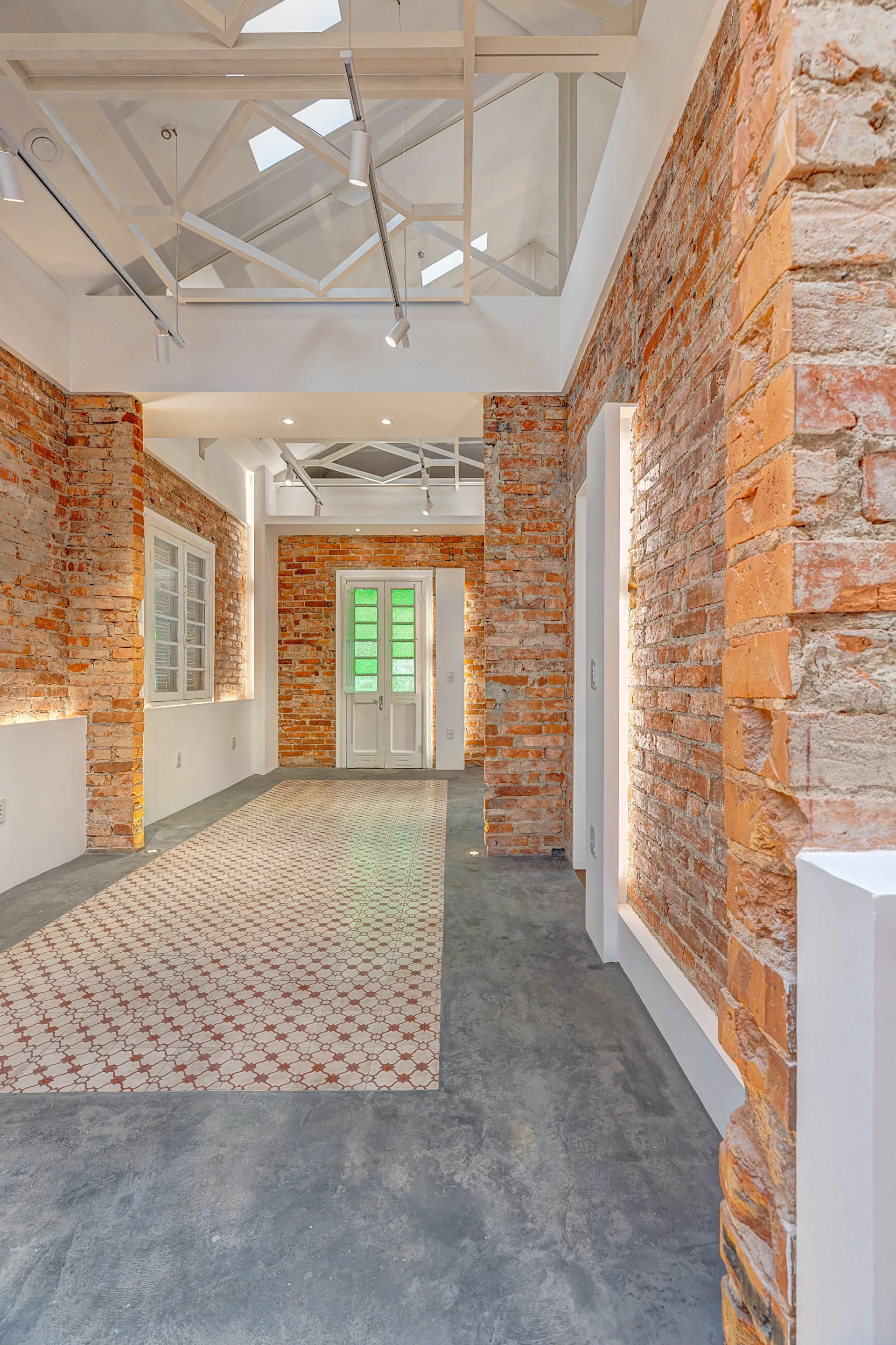 A long hallway features exposed red brick walls, a patterned tile runner set in a grey concrete floor, and white geometric ceiling trusses.