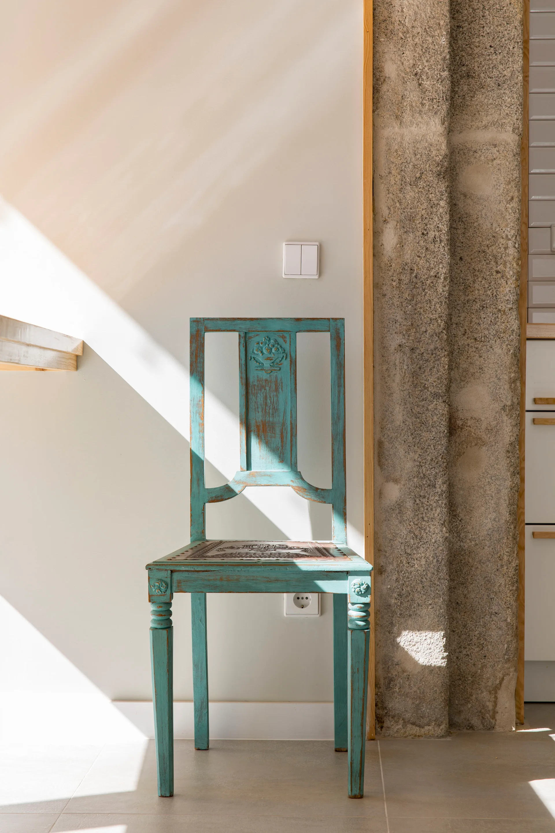 A weathered turquoise wooden chair stands in the sunlight against a white wall and a textured stone pillar.