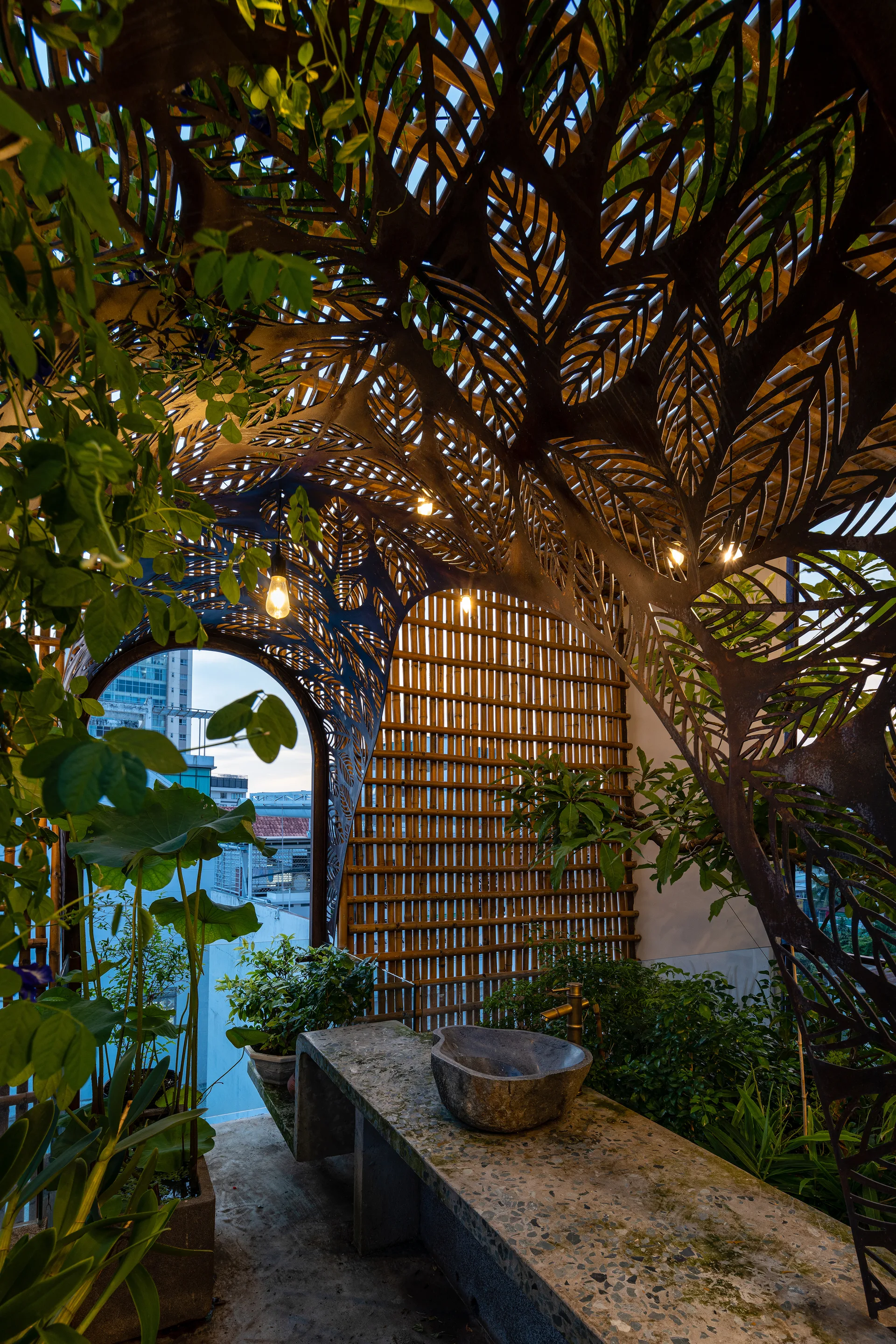 An outdoor balcony vanity features a stone sink on a concrete counter beneath a leaf-patterned metal canopy and a bamboo privacy wall.