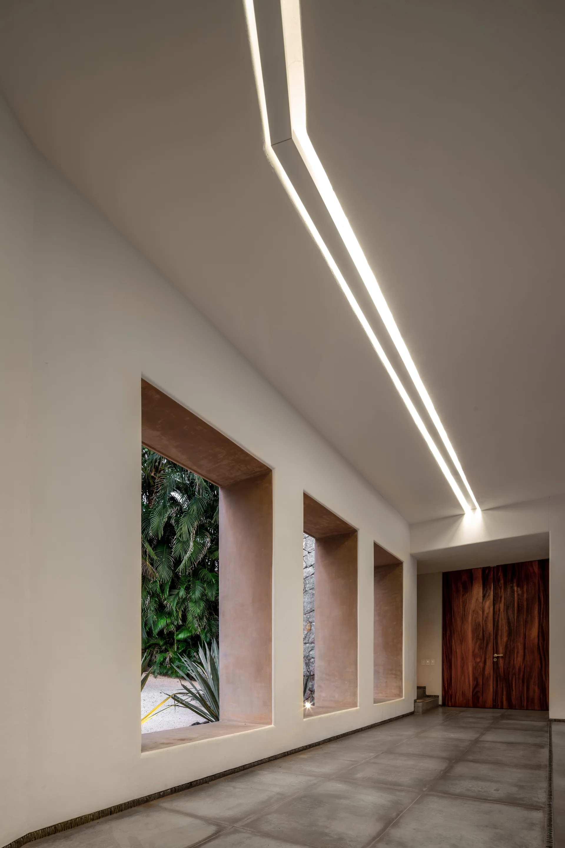 A minimalist hallway features concrete tile floors, a long recessed linear ceiling light, deep-set square window openings, and a rich wood door.