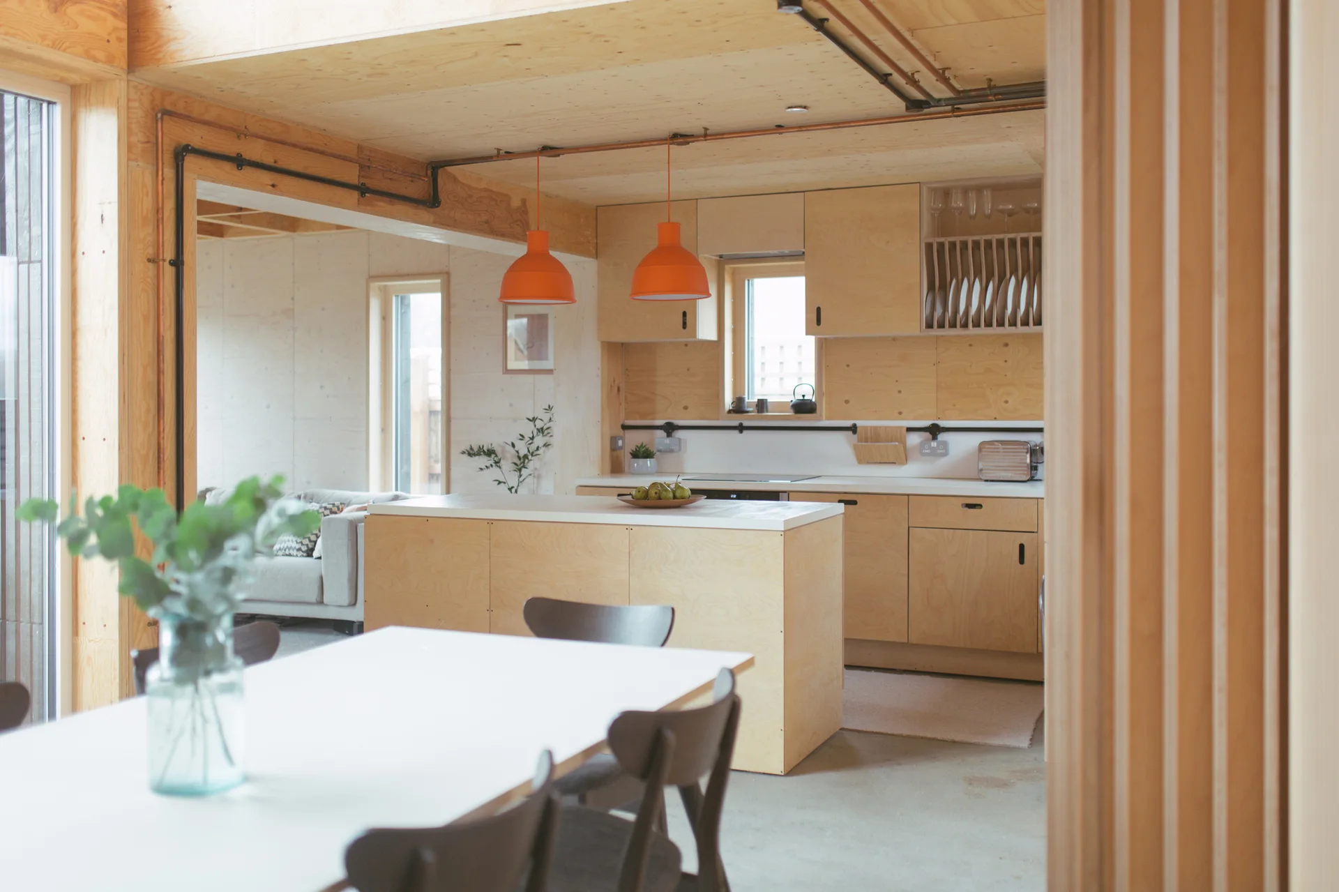 A modern open-plan kitchen featuring light plywood cabinets, a central island, and two hanging orange pendant lights.