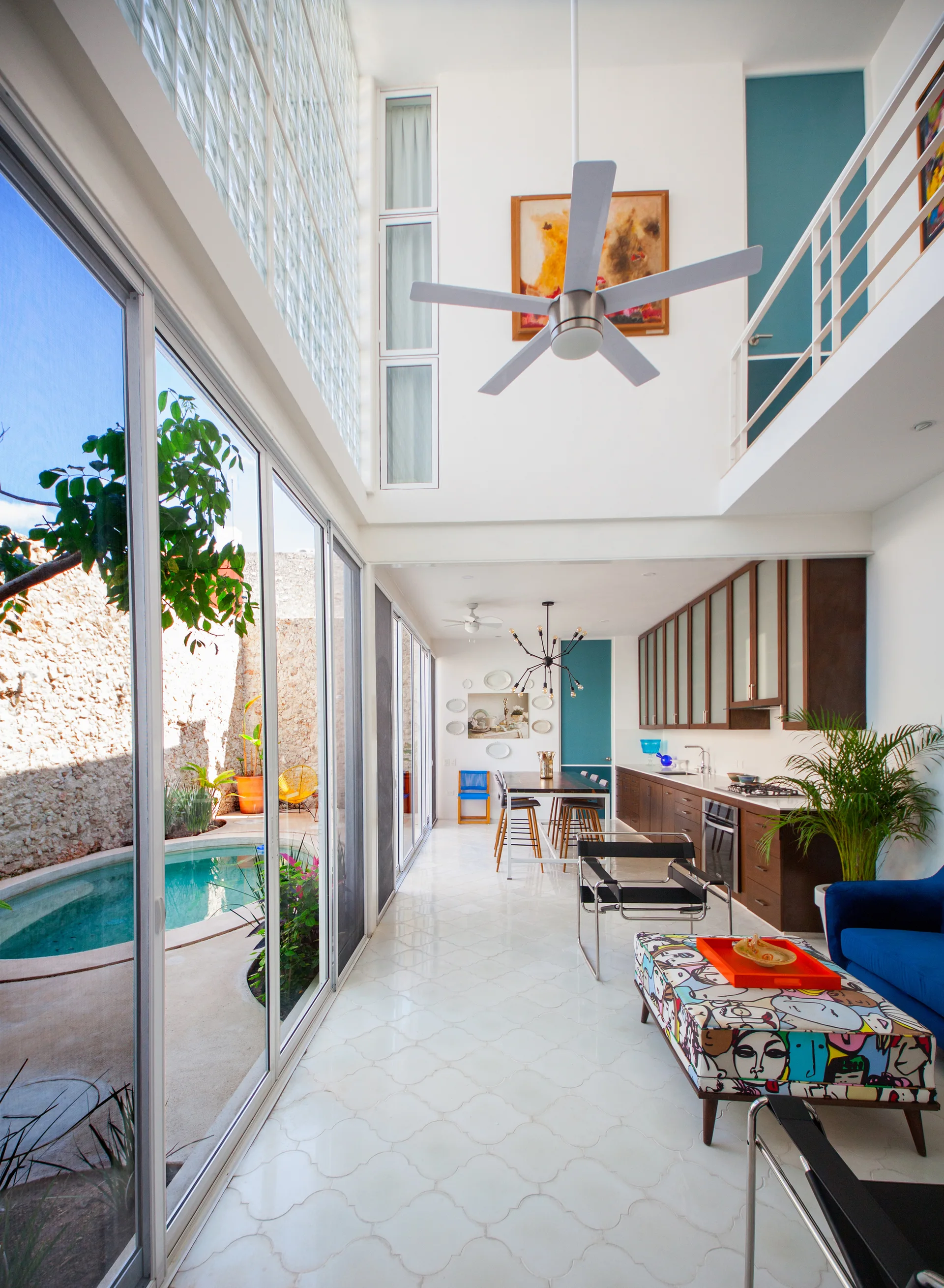 A modern double-height interior with a kitchen and living area next to a glass wall overlooking a swimming pool.