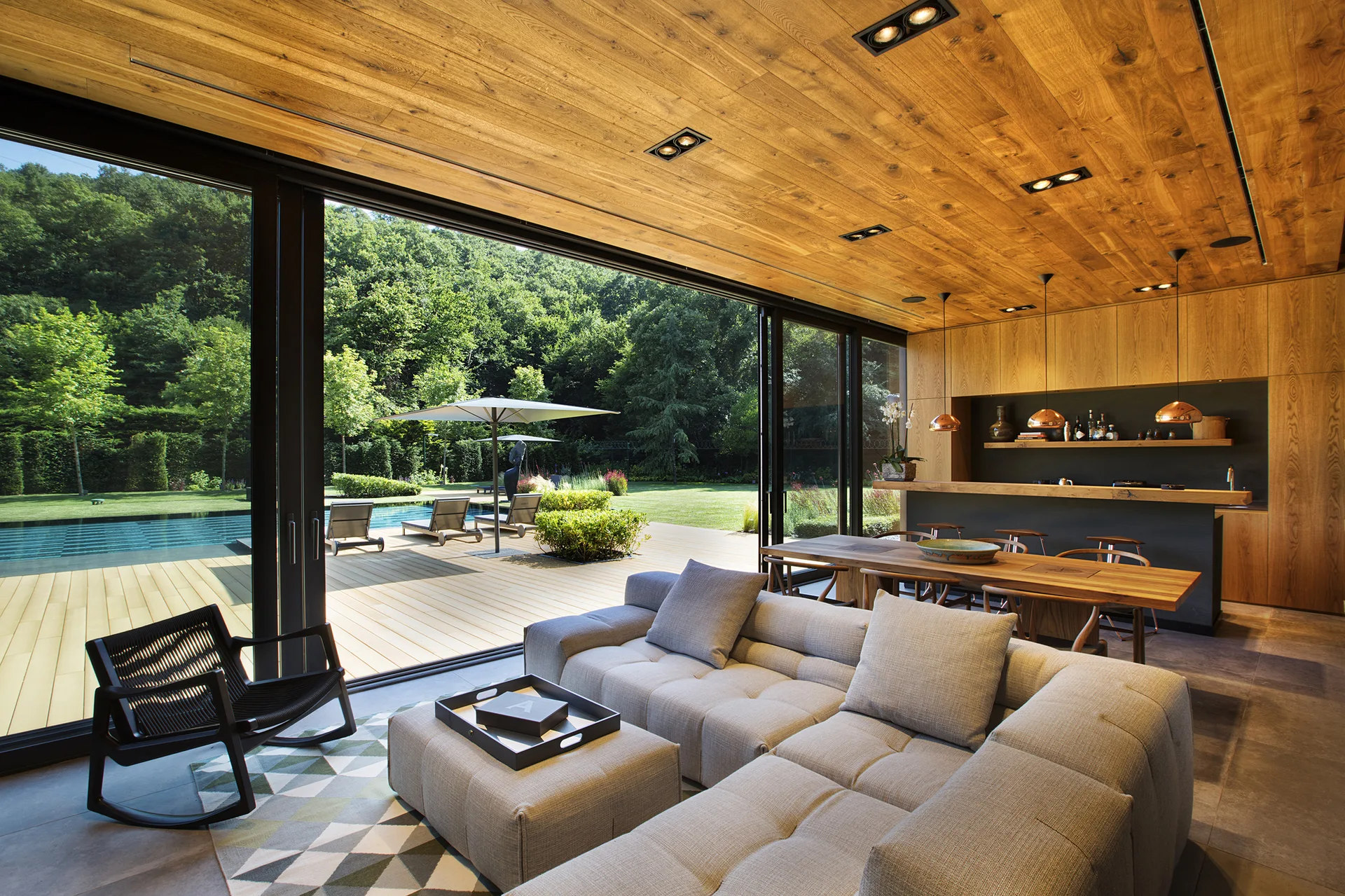 A modern open-concept living area features a grey sectional sofa, wood plank ceiling, and large sliding glass doors opening to an outdoor pool deck.