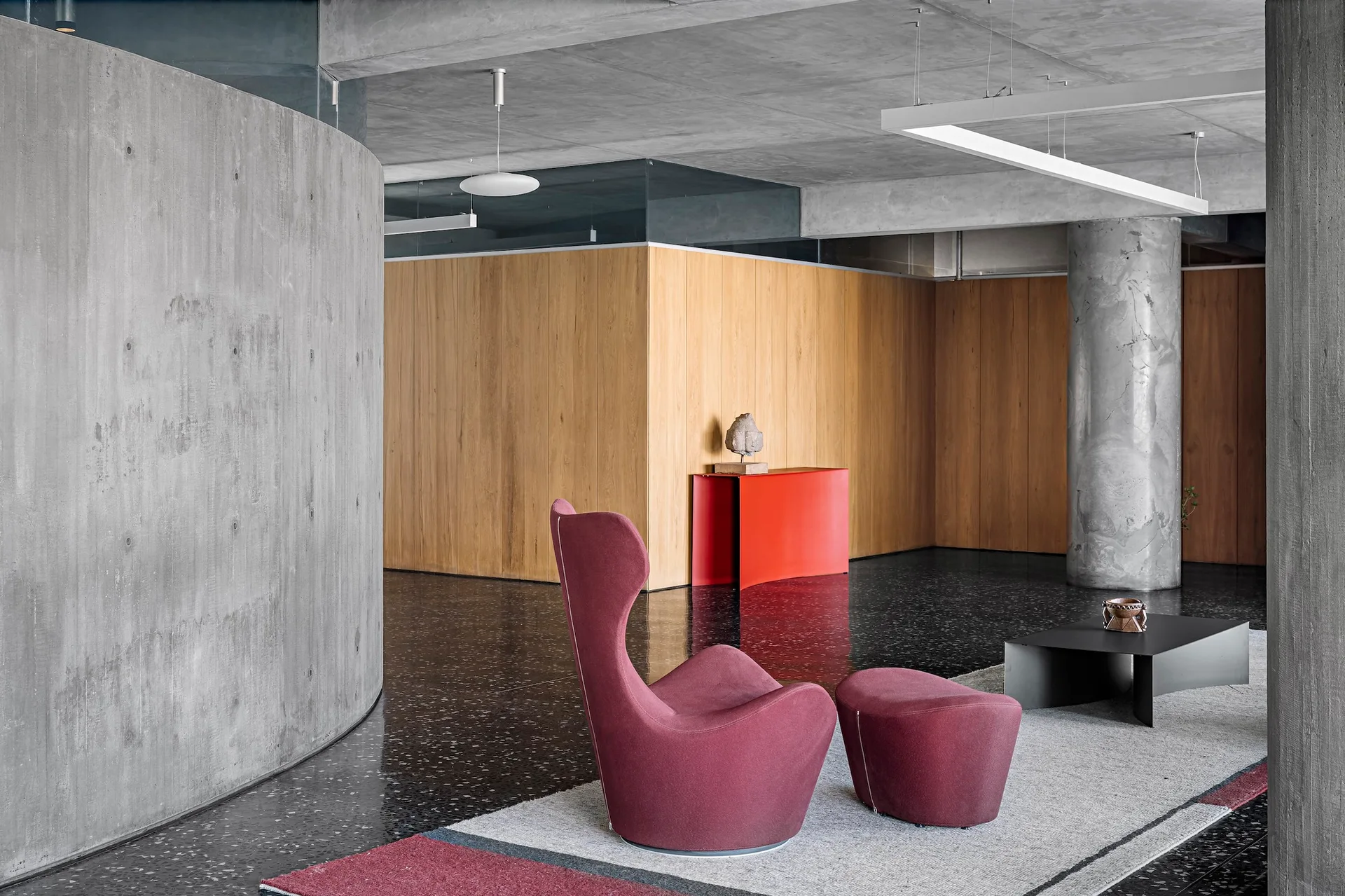 A modern living space with a curved concrete partition, a burgundy wing chair with matching ottoman, warm wood paneling, and a polished dark terrazzo floor.