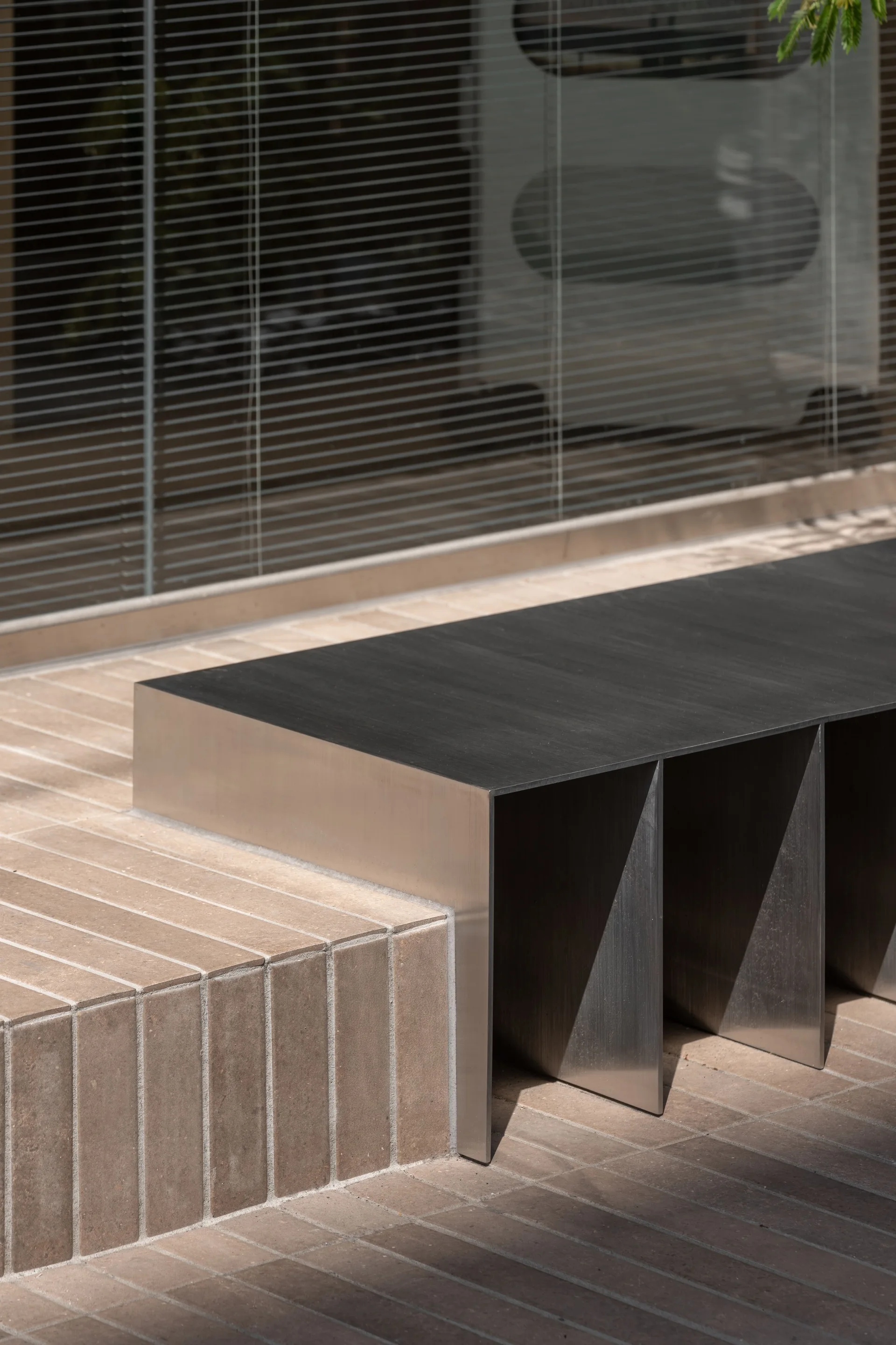 A modern brushed metal bench is integrated into a brown paved brick step in front of a large window with horizontal blinds.
