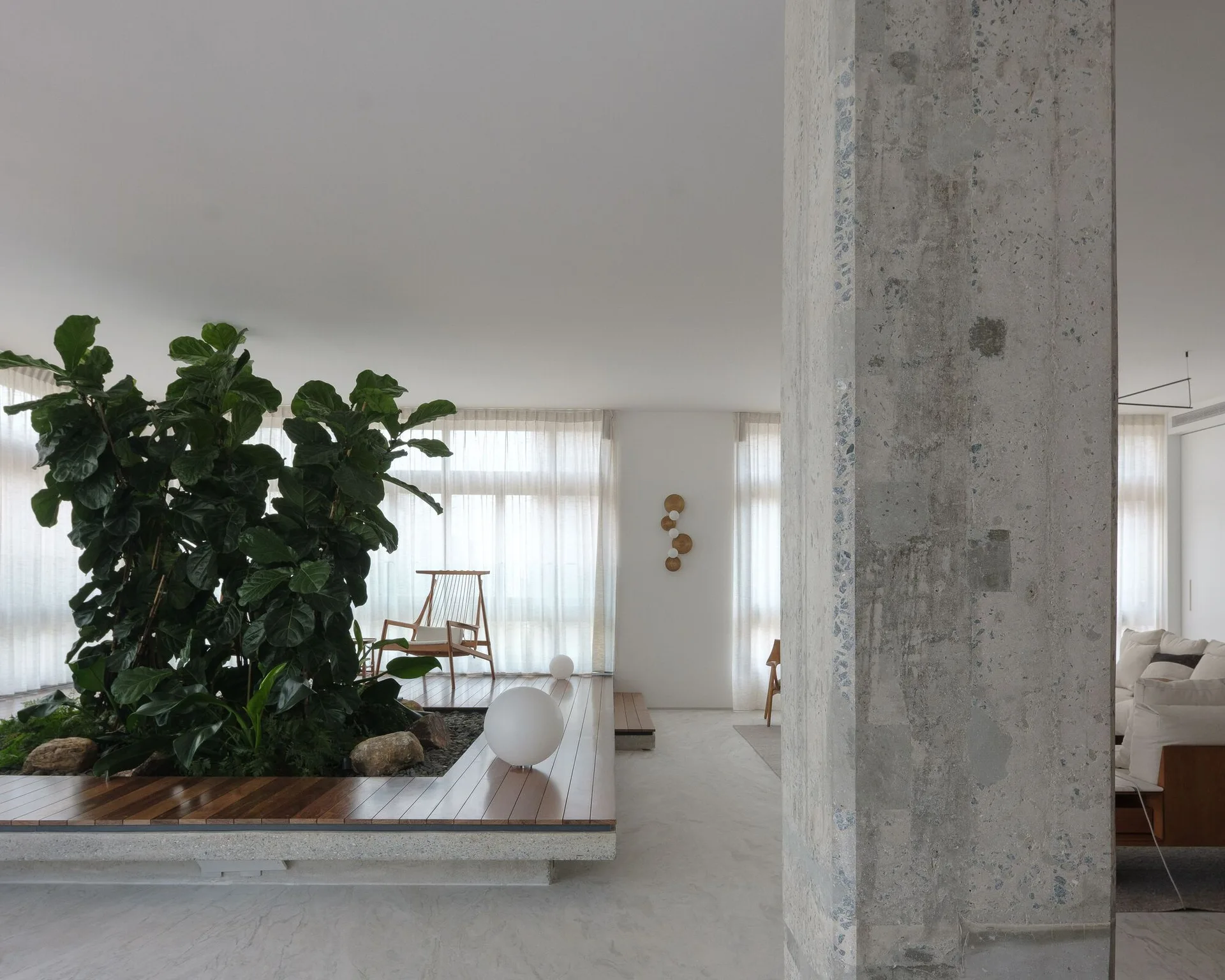 A minimalist living room featuring a large concrete column in the foreground, a raised wooden platform with an indoor garden, and sheer white curtains.