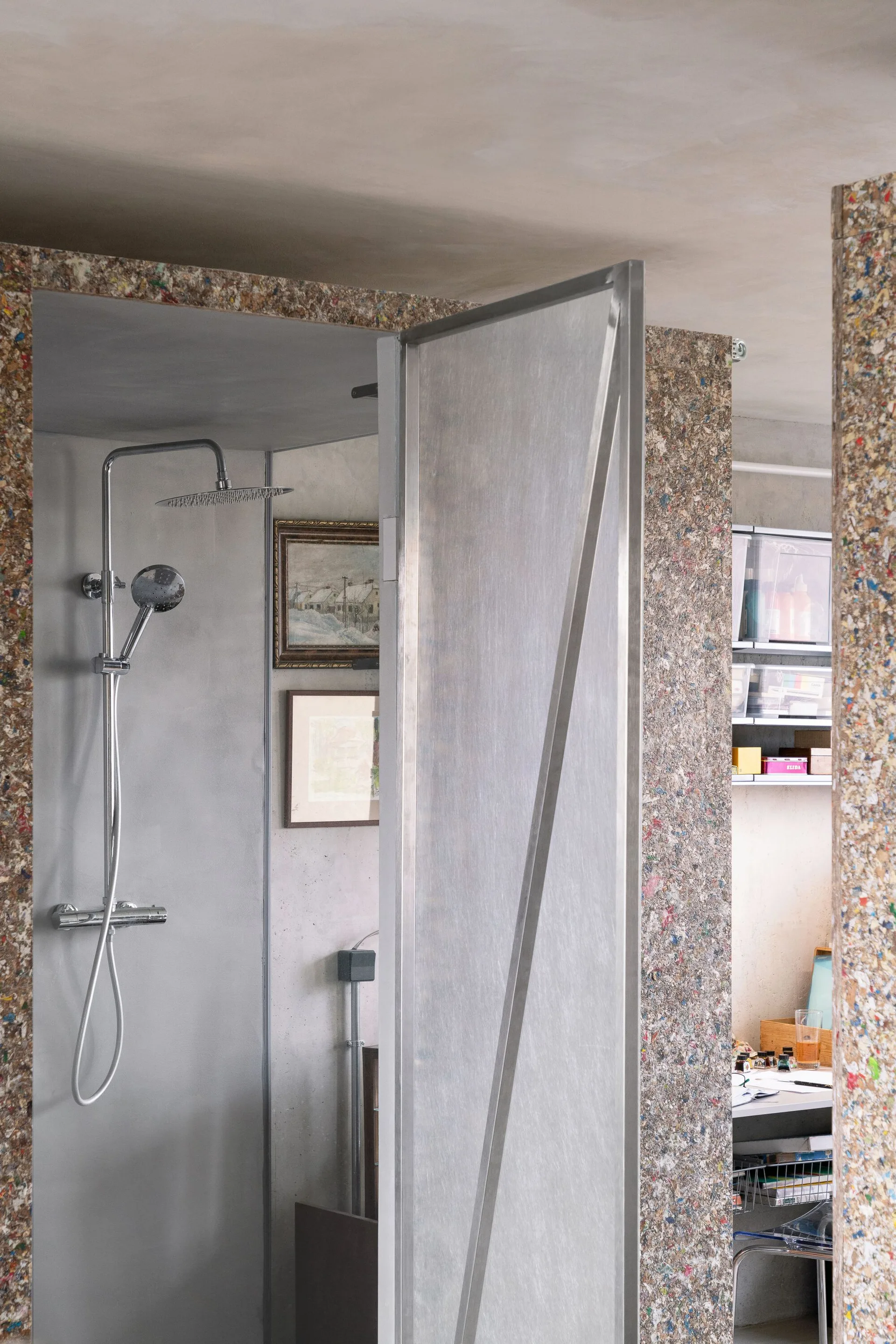 A modern shower with a chrome fixture and metal door is surrounded by multicolored speckled recycled composite wall panels.