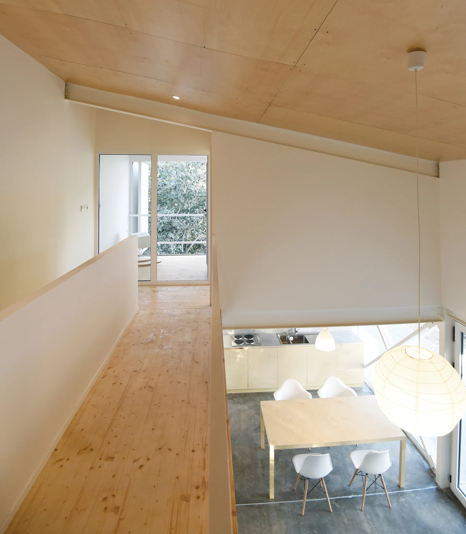 An elevated view from a light wood mezzanine looking down at a dining table, white chairs, and a paper pendant light on a concrete floor.