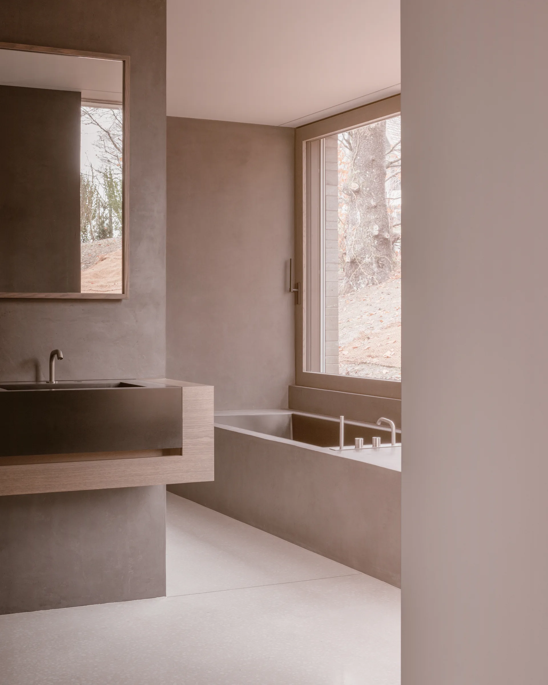 A minimalist bathroom featuring a concrete-finished bathtub, a cantilevered wooden vanity with a dark sink, and a large window looking out at trees.