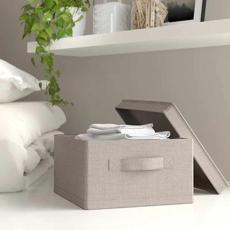 A beige woven fabric storage bin with its lid leaning beside it holds folded white linens beneath a white floating wall shelf.