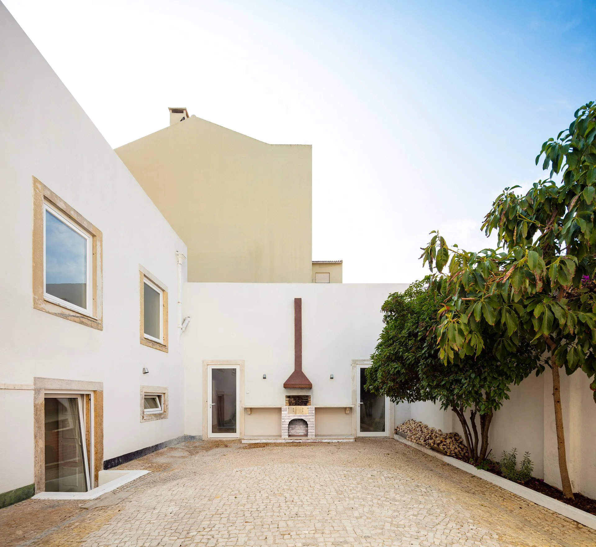 A residential white stucco courtyard features a built-in masonry barbecue, cobblestone paving, and a green tree against a high wall.