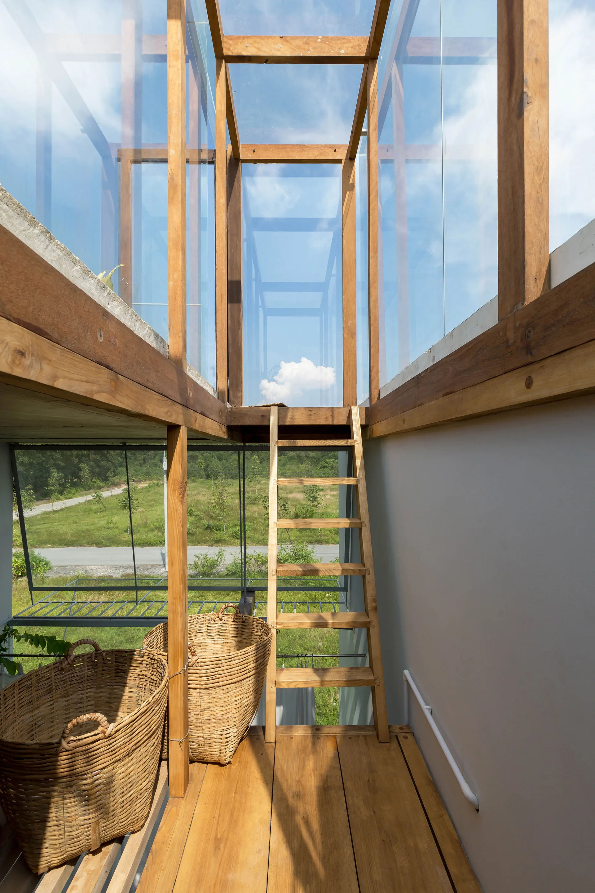 A wooden ladder sits on a plank floor next to large wicker baskets, leading up to an enclosed glass corridor with exposed timber framing.