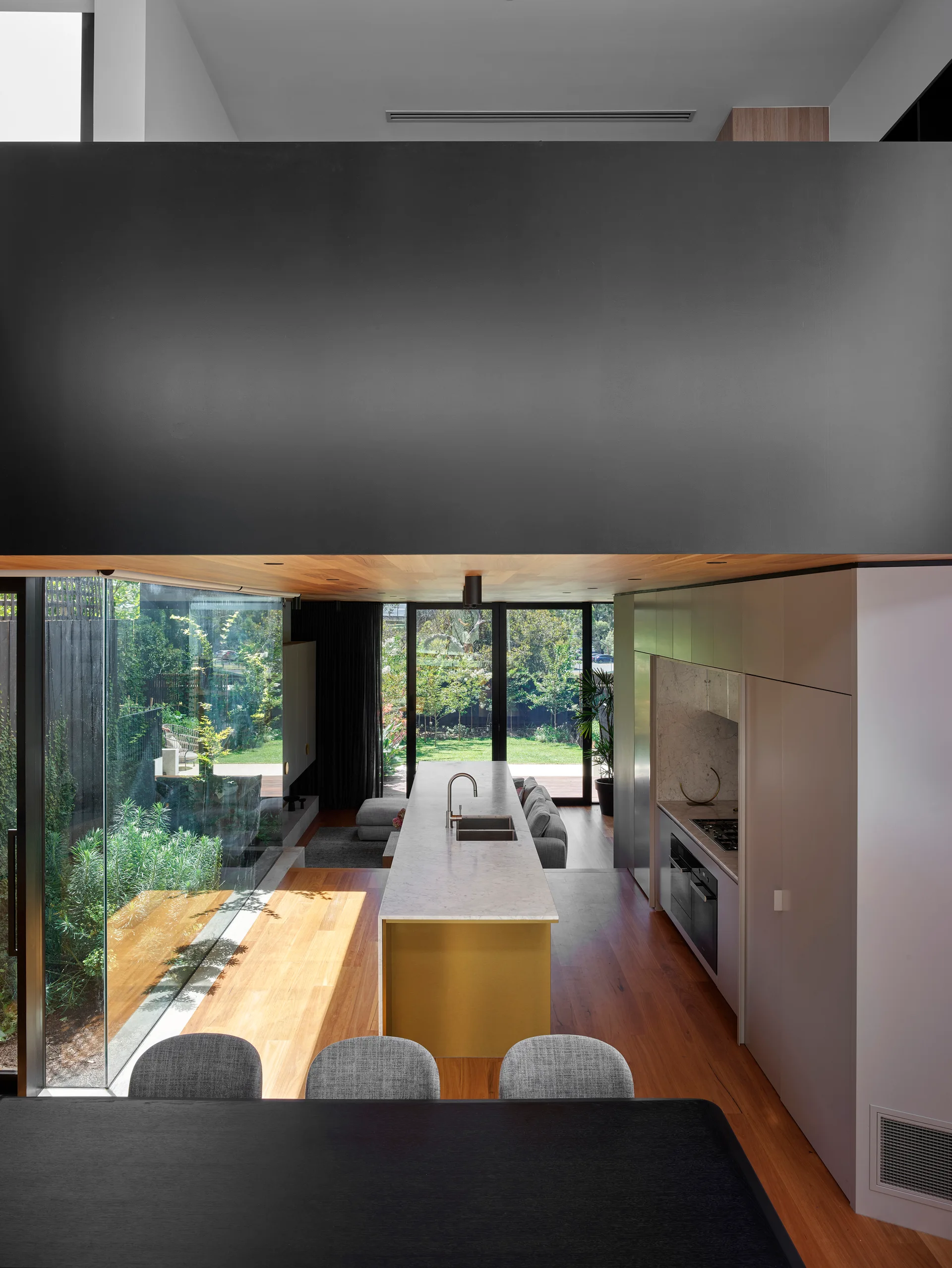 A high-angle view of a minimalist kitchen featuring a long stone-topped island with a gold metallic base, surrounded by wood floors and large windows.