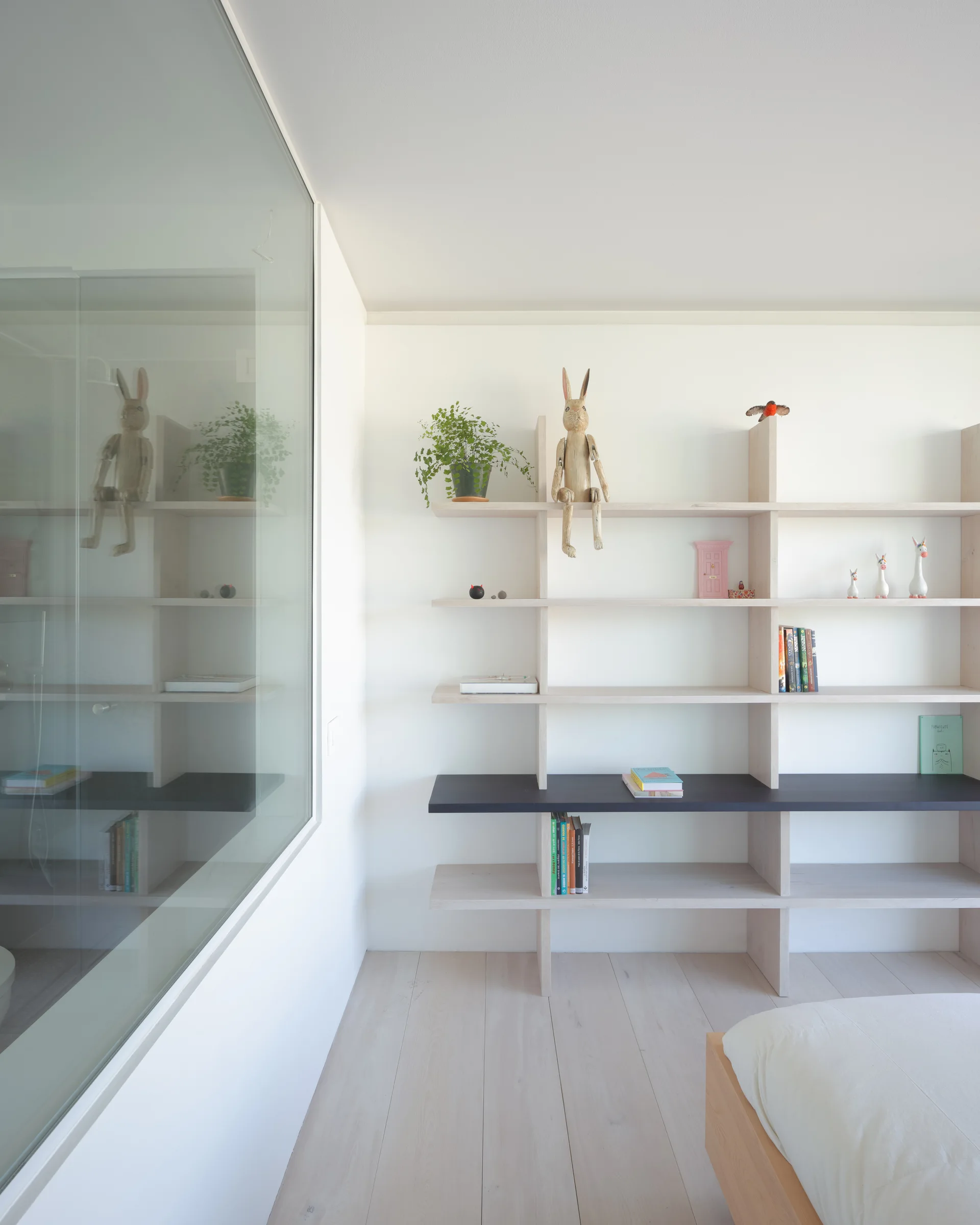 A minimalist bedroom featuring light wood flooring, a glass partition wall, and a large wooden shelving unit styled with plants and figurines.