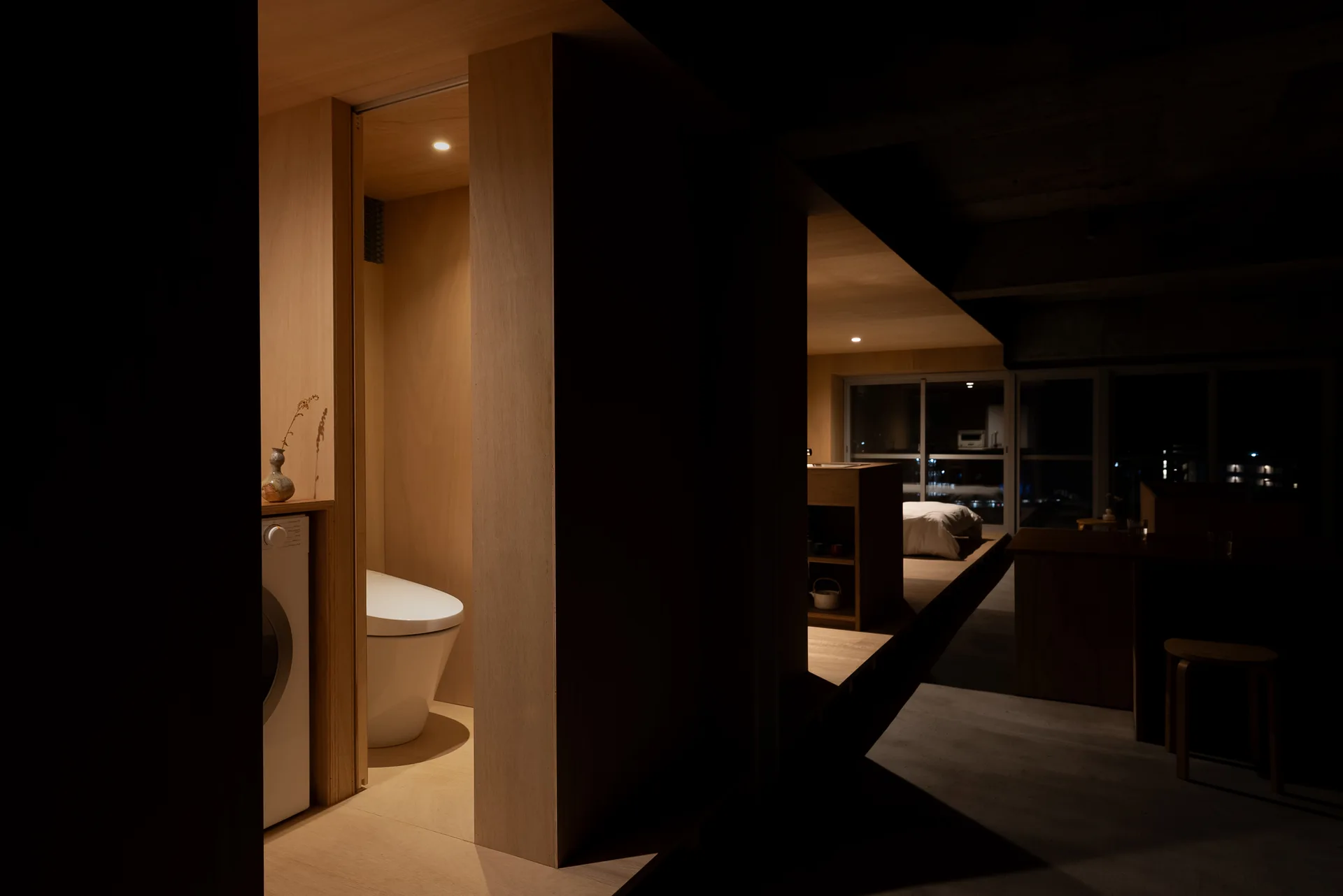 A dimly lit minimalist interior showing a plywood-paneled bathroom with a toilet and washer next to an open bedroom on a raised platform.