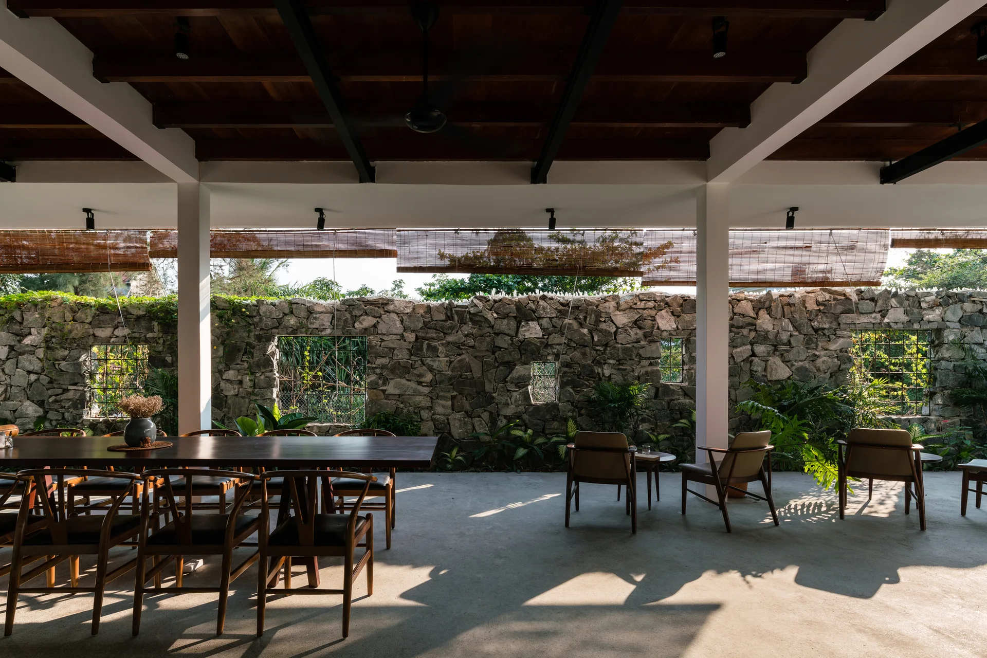 An open-air patio features a dark wood dining table with chairs and lounge seating arranged on concrete floors in front of a rustic stone wall.