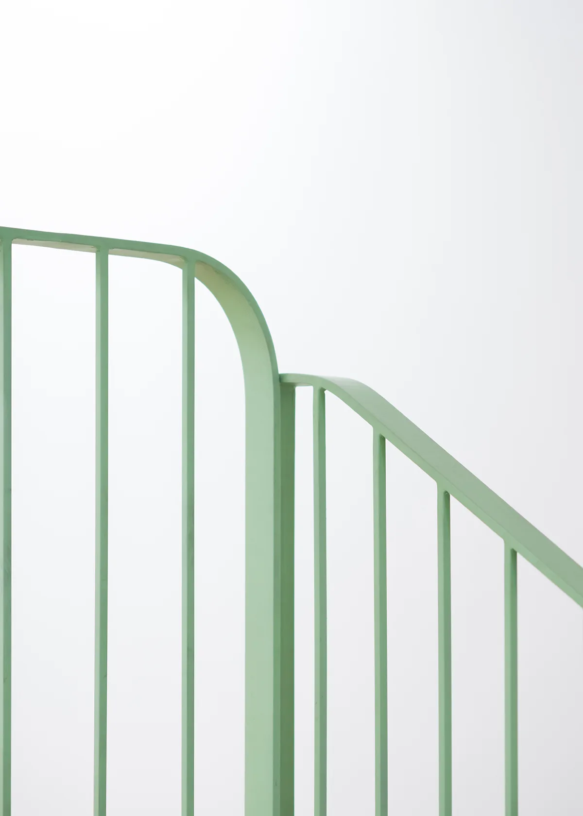 A close-up of a pale green metal staircase railing with rounded corners and vertical slats against a solid white wall.