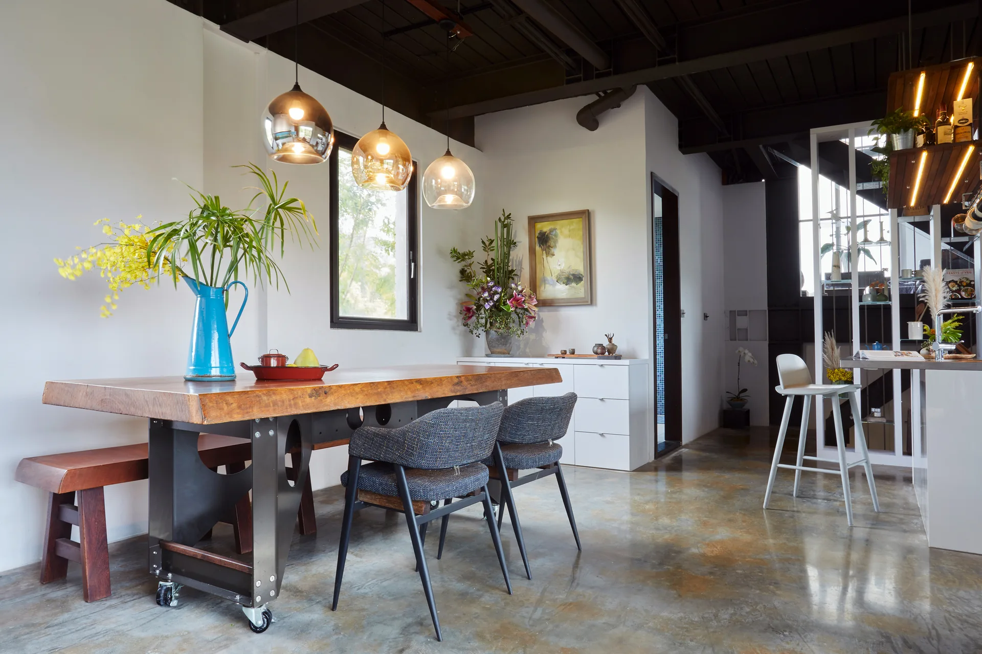 An industrial dining room features a live-edge wood table, glass pendant lights, and a polished concrete floor under exposed ceiling beams.