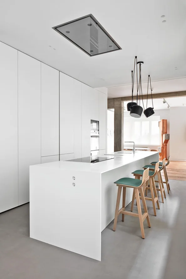 A minimalist white kitchen features a long white island with wooden bar stools, handleless floor-to-ceiling cabinets, and black pendant lights.
