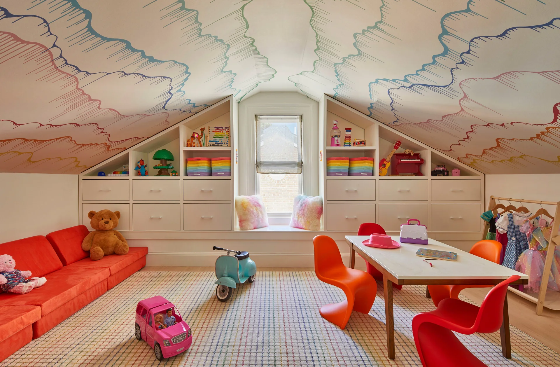 A children's attic playroom with colorful cloud wallpaper on the sloped ceiling, built-in white cabinets, a window seat, and bright orange furniture.
