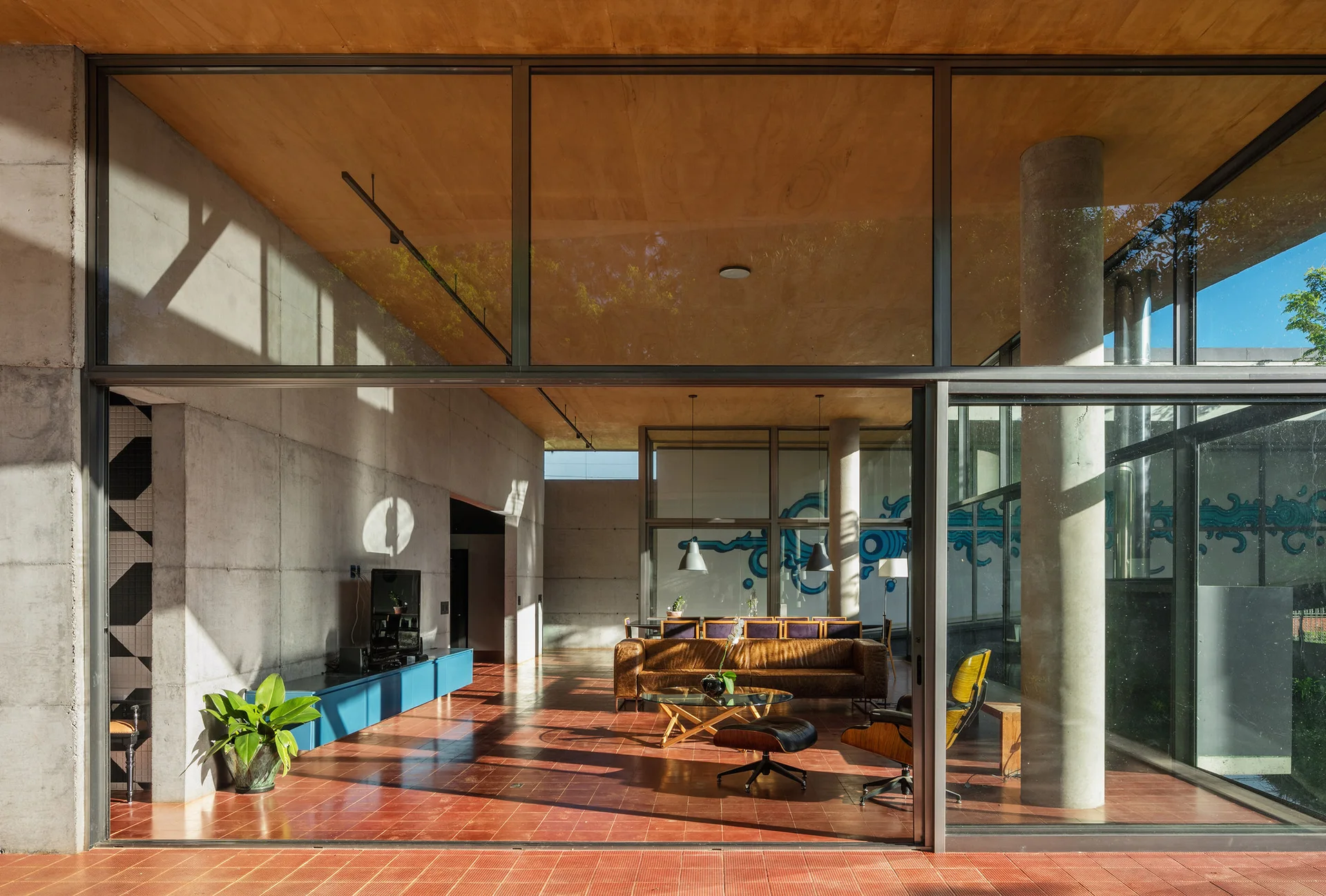A spacious open-plan living room displays raw concrete walls, a warm plywood ceiling, red tile floors, a leather sofa, and a blue media console.