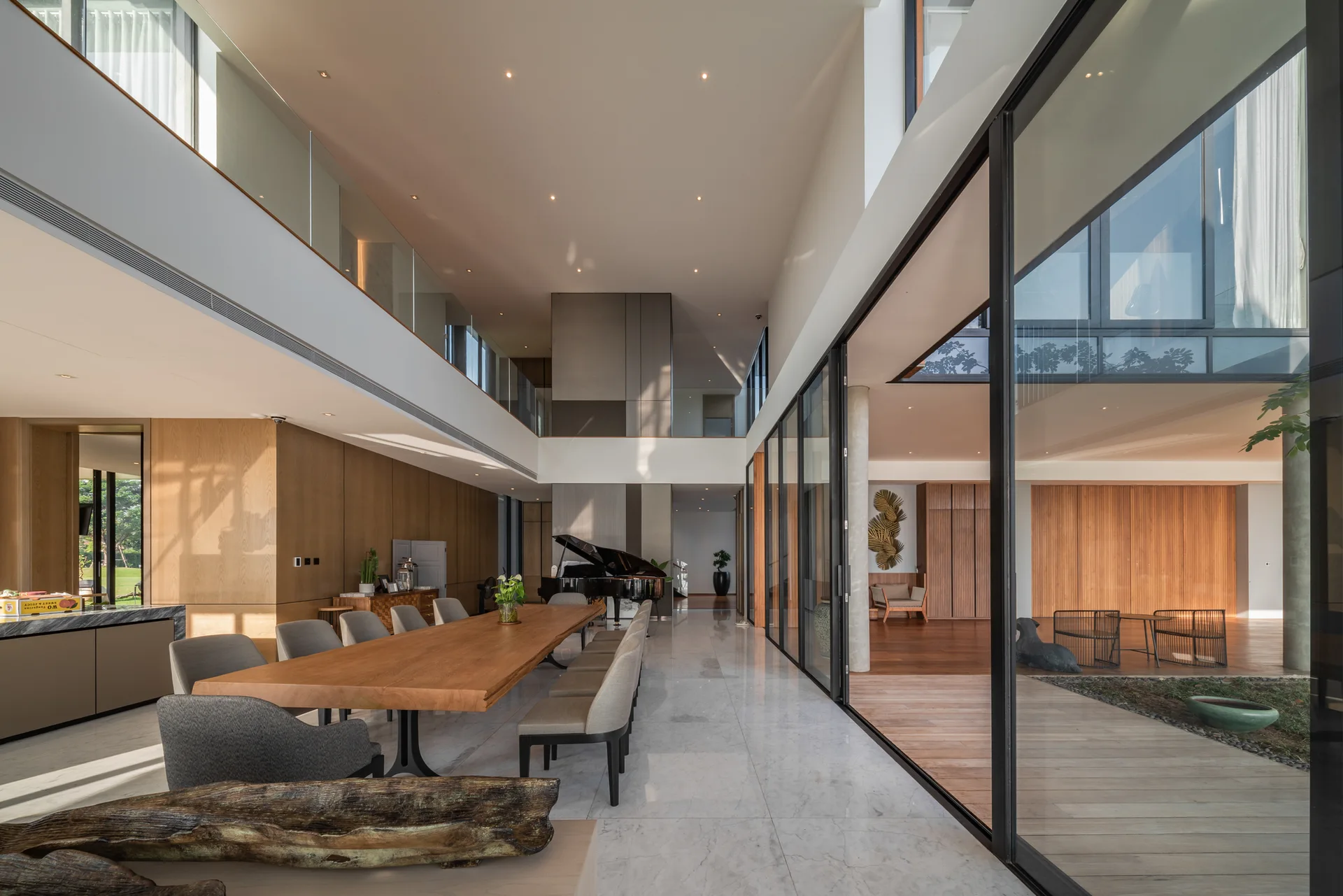 A long wooden dining table with grey chairs sits in a modern open-plan room with polished marble floors, a mezzanine, and glass courtyard walls.
